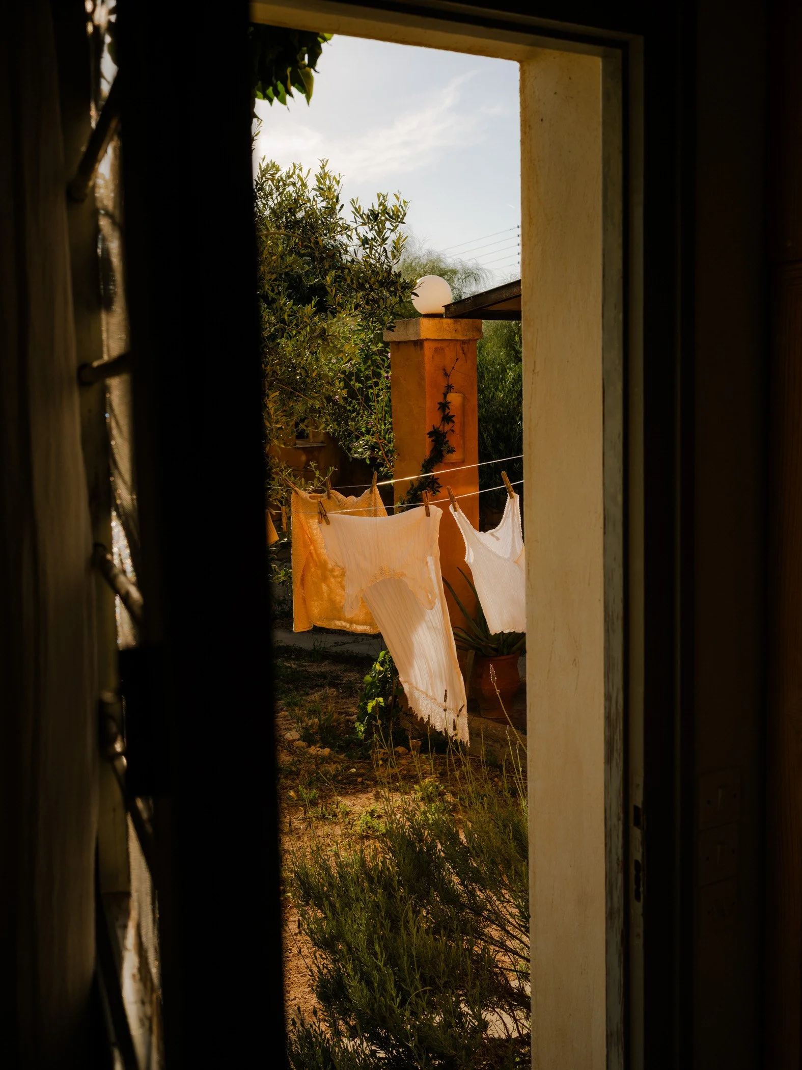 A view through a window shows laundry hanging outside on a clothesline, with greenery and trees in the background, and a yellow outdoor lamp on a post.