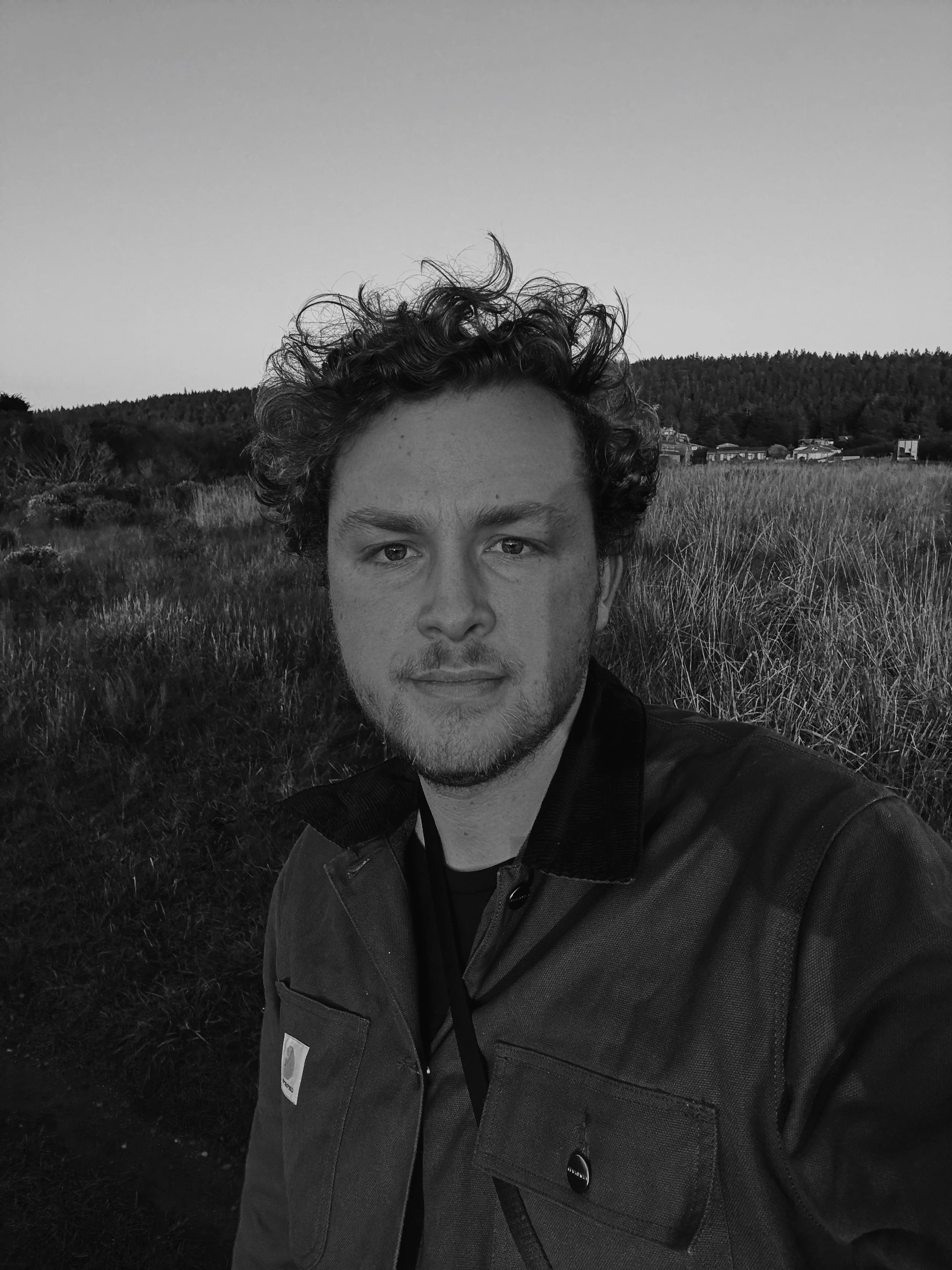 A man taking a selfie outdoors in a field with grass, with a forested hill and houses in the background, in black and white.