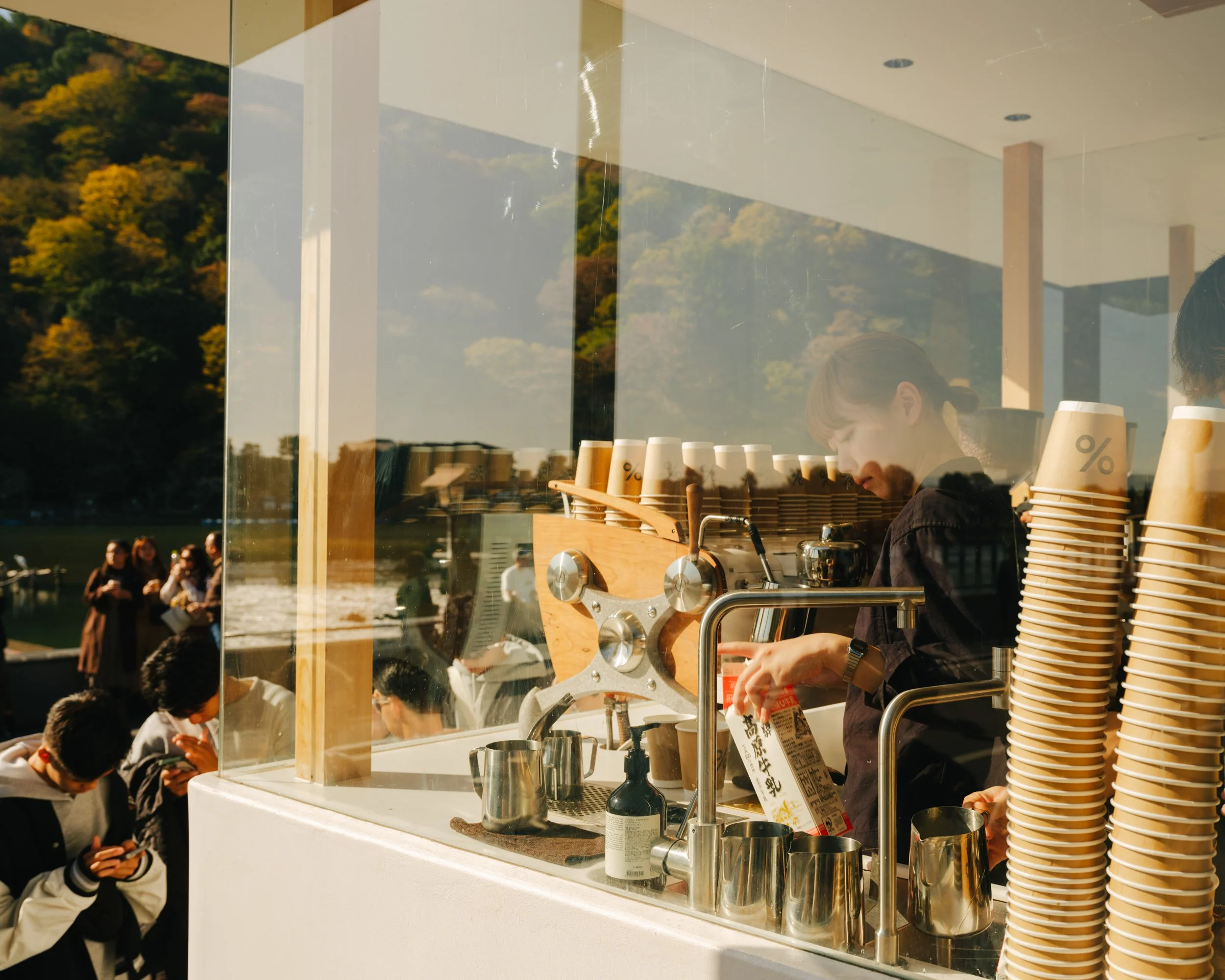 People lined up at a coffee stand outdoors with trees and a park in the background, seen through a glass window.