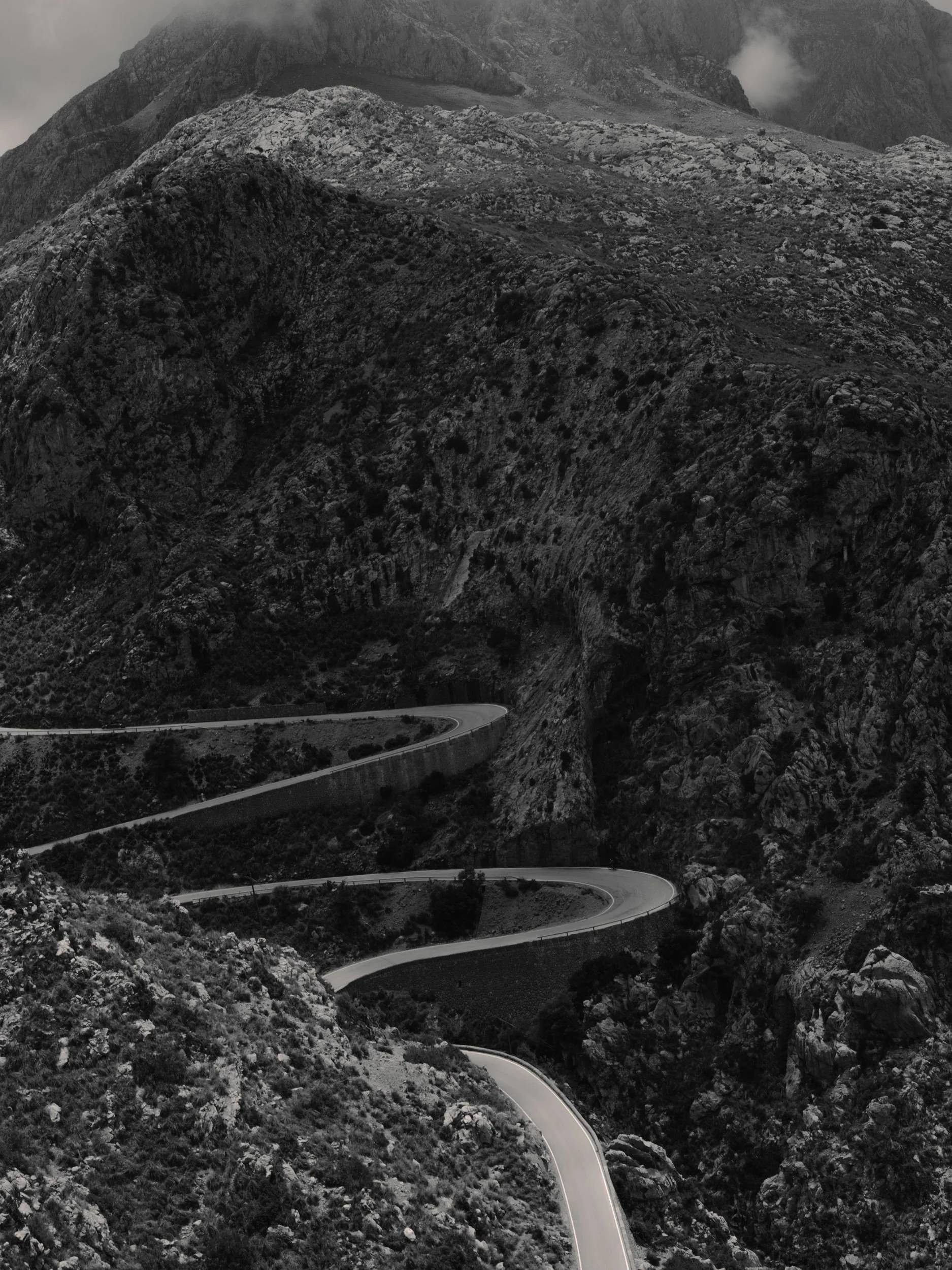 Black and white photo of a winding mountain road with sharp curves, surrounded by rugged terrain and rocky slopes.