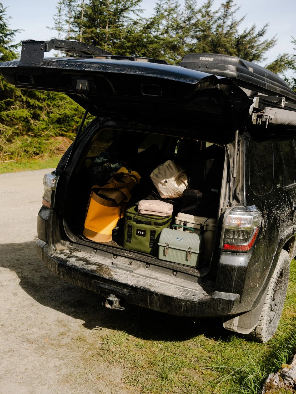 Open SUV trunk filled with camping gear, including bags, coolers, and a small storage box, on a dirt road surrounded by greenery and trees.