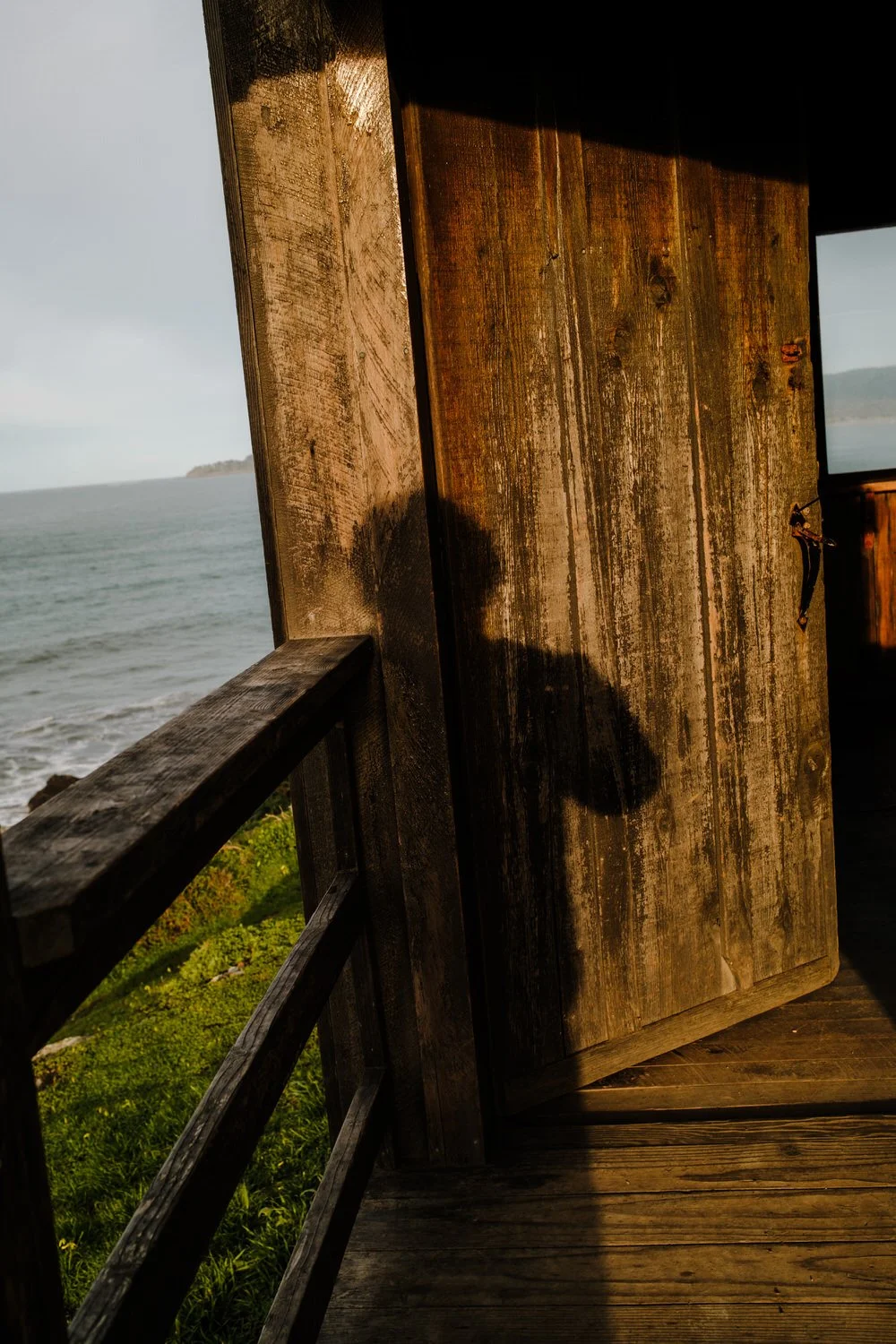 Sunlight casting a shadow of a person on a wooden door of a coastal cabin, overlooking the ocean with islands in the distance.