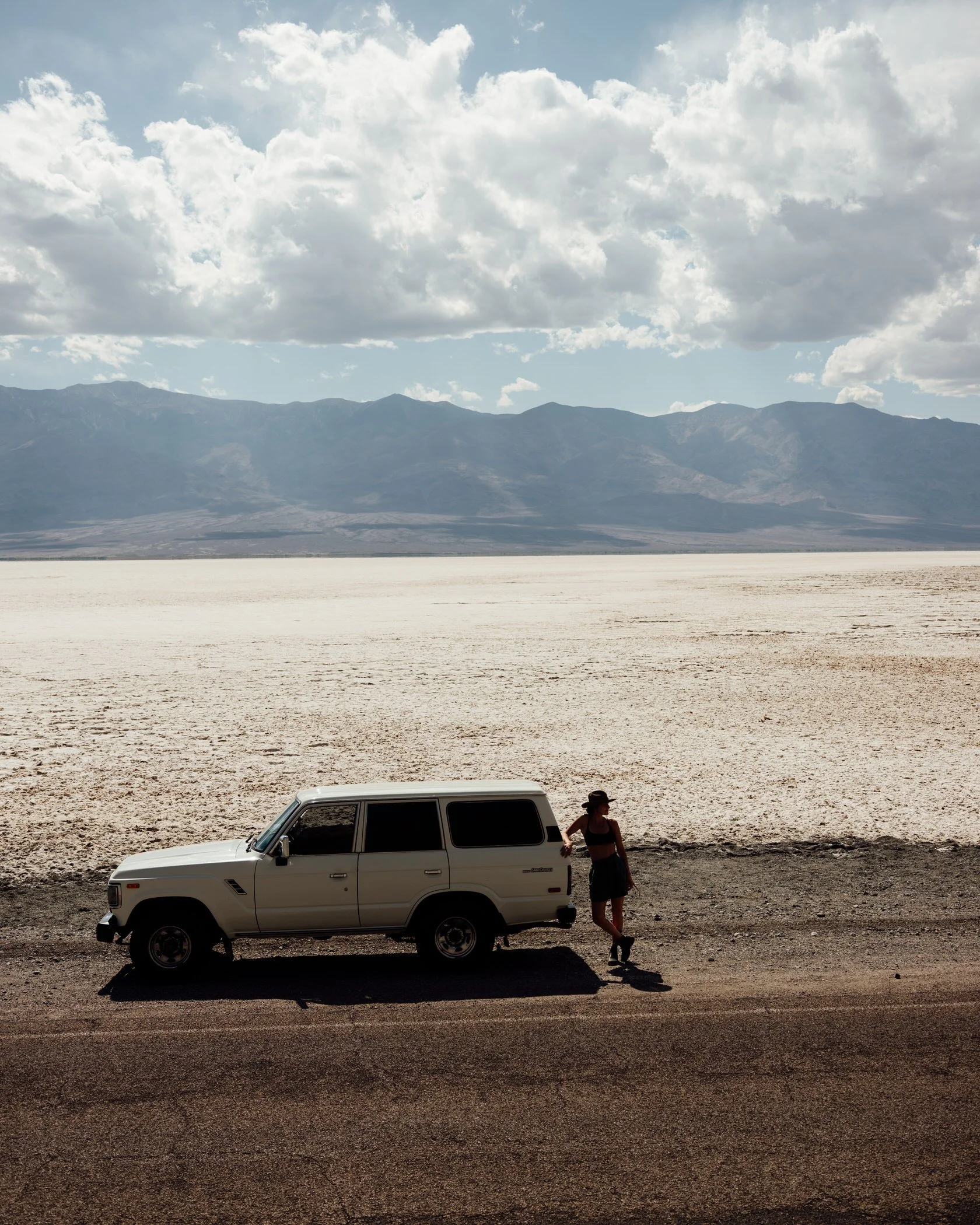 A woman standing next to a white SUV parked by the side of a road in a desert with mountain ranges in the background, under cloudy skies.