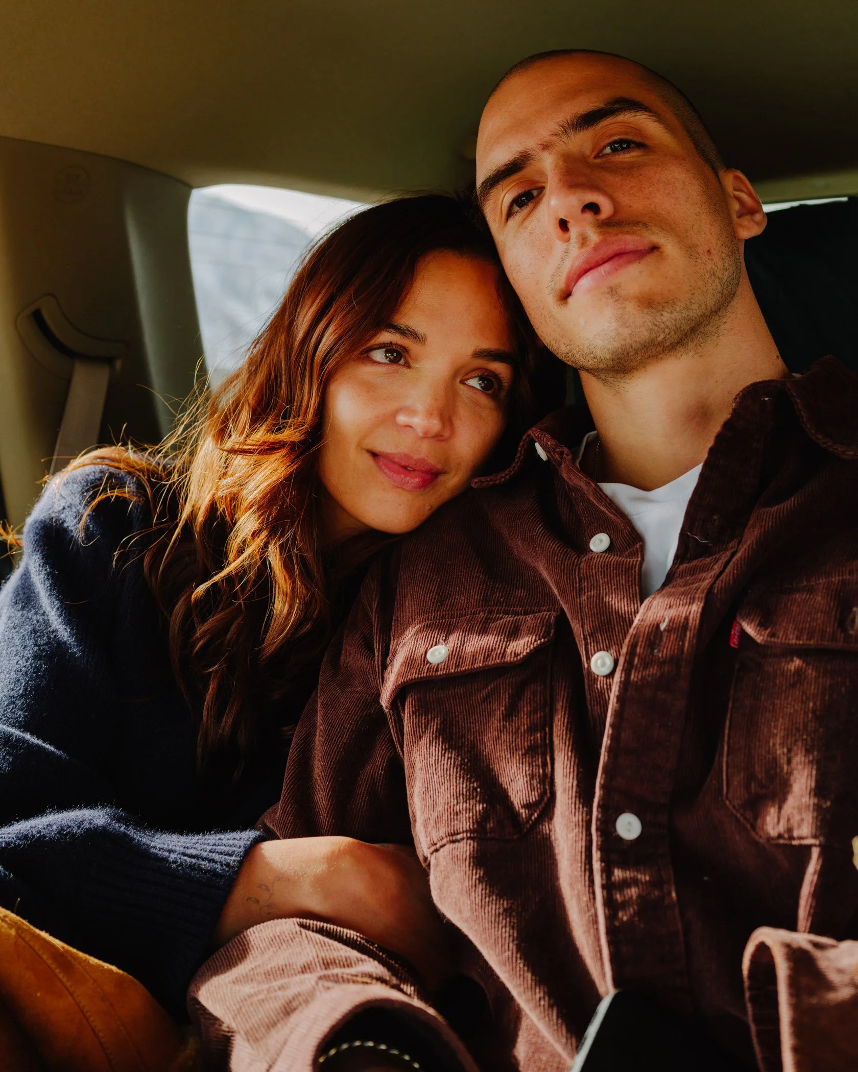 A young man and woman taking a selfie together in a vehicle. The woman has long brown hair and the man has a shaved head. They are close together and smiling softly.