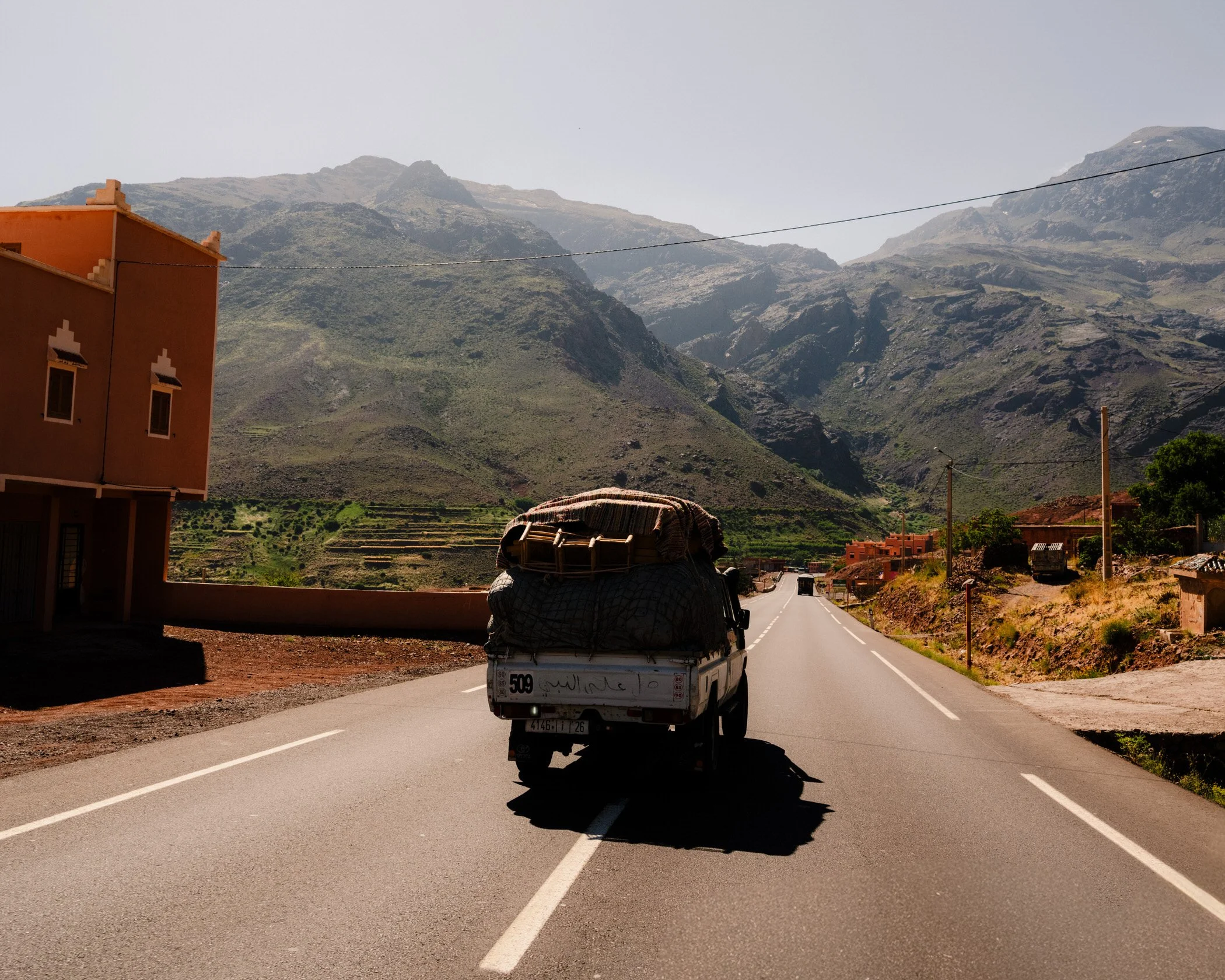 A pickup truck driving down a two-lane road with a mountainous landscape in the background. The truck is carrying loads covered with a blanket, and a building with an orange facade is on the left side of the road.