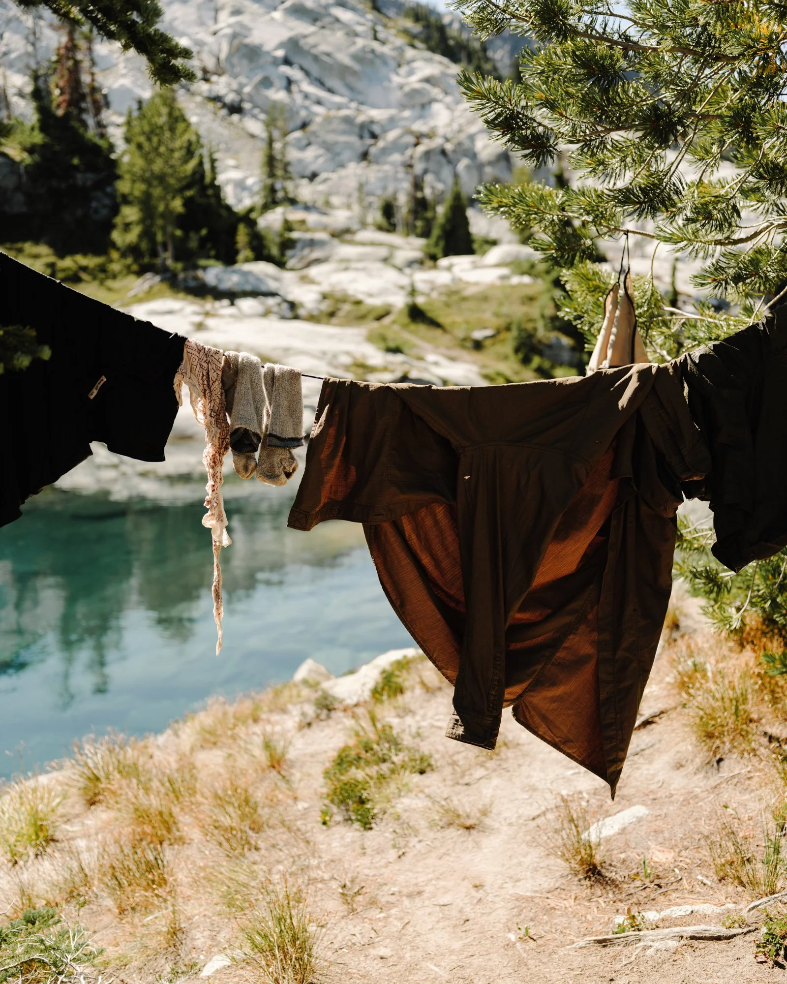 Clothes hanging on a line between trees near a mountain lake, with rocky mountains and evergreen trees in the background.
