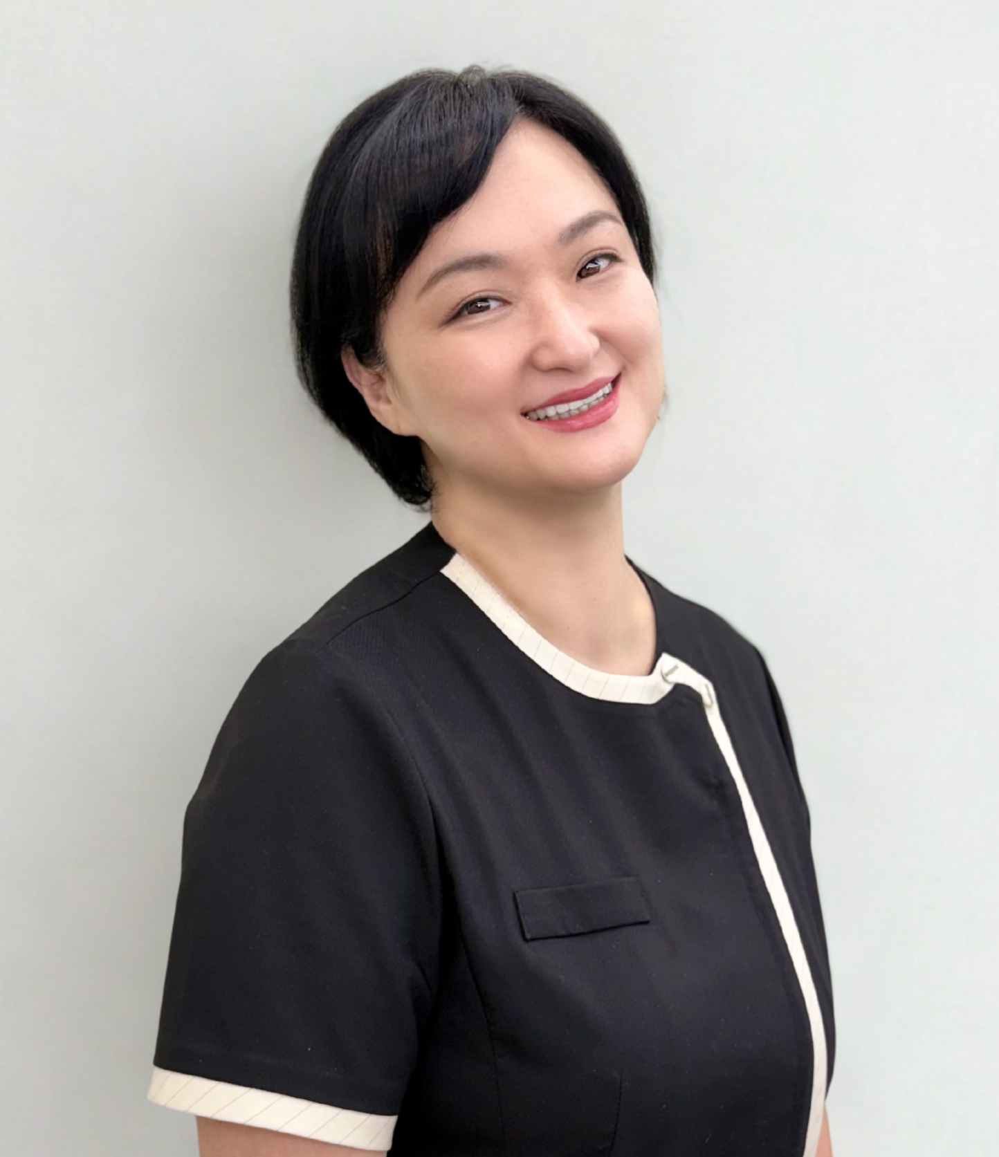 A woman with short black hair, smiling, wearing a black dress with a white trim, standing against a plain light background.