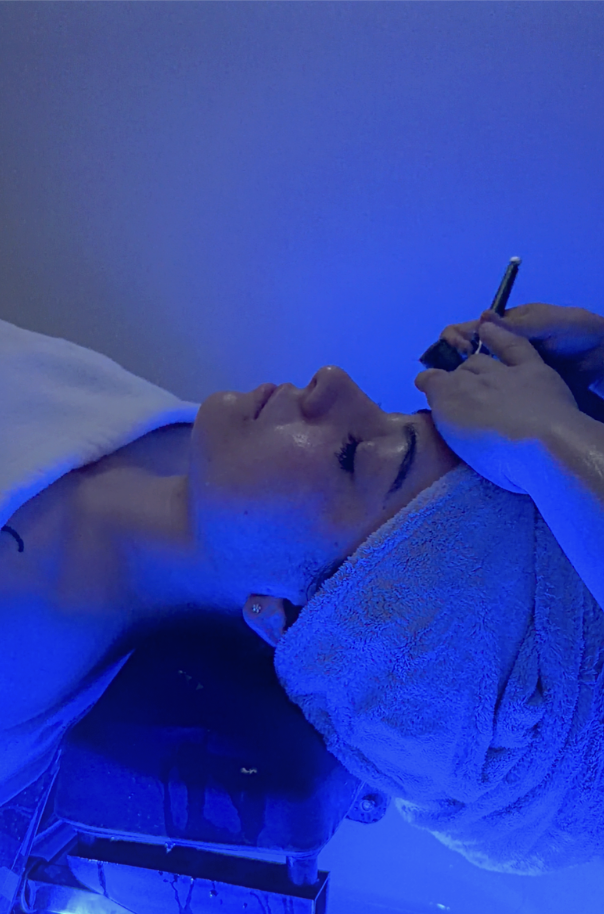 A woman receiving a facial treatment while lying down with her eyes closed, wrapped in a towel, in a spa or skincare clinic, with a person applying a skincare product on her face under blue lighting.