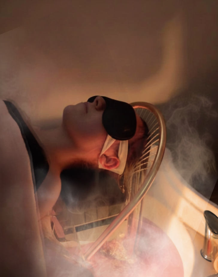 Person lying on a massage table with face covered by a black eye mask, surrounded by steam or mist, with a copper or bronze decorative railing nearby.