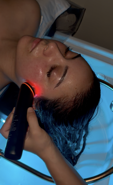 A woman receives a facial treatment with a handheld device emitting red light while lying in a reclining treatment bed, with her eyes closed and wet hair.