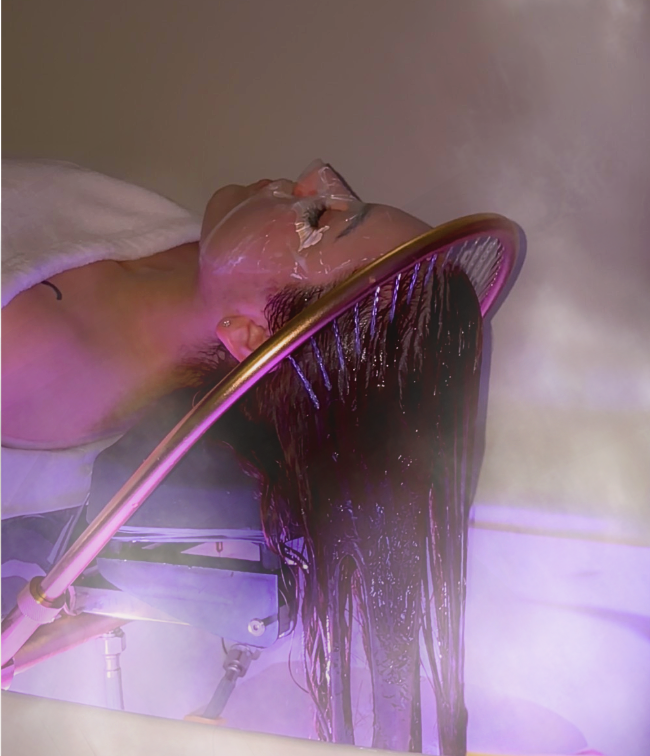 A woman receiving a cryotherapy or similar treatment with her eyes closed, and her hair hanging downward, while covered with a transparent mask and surrounded by vapor.