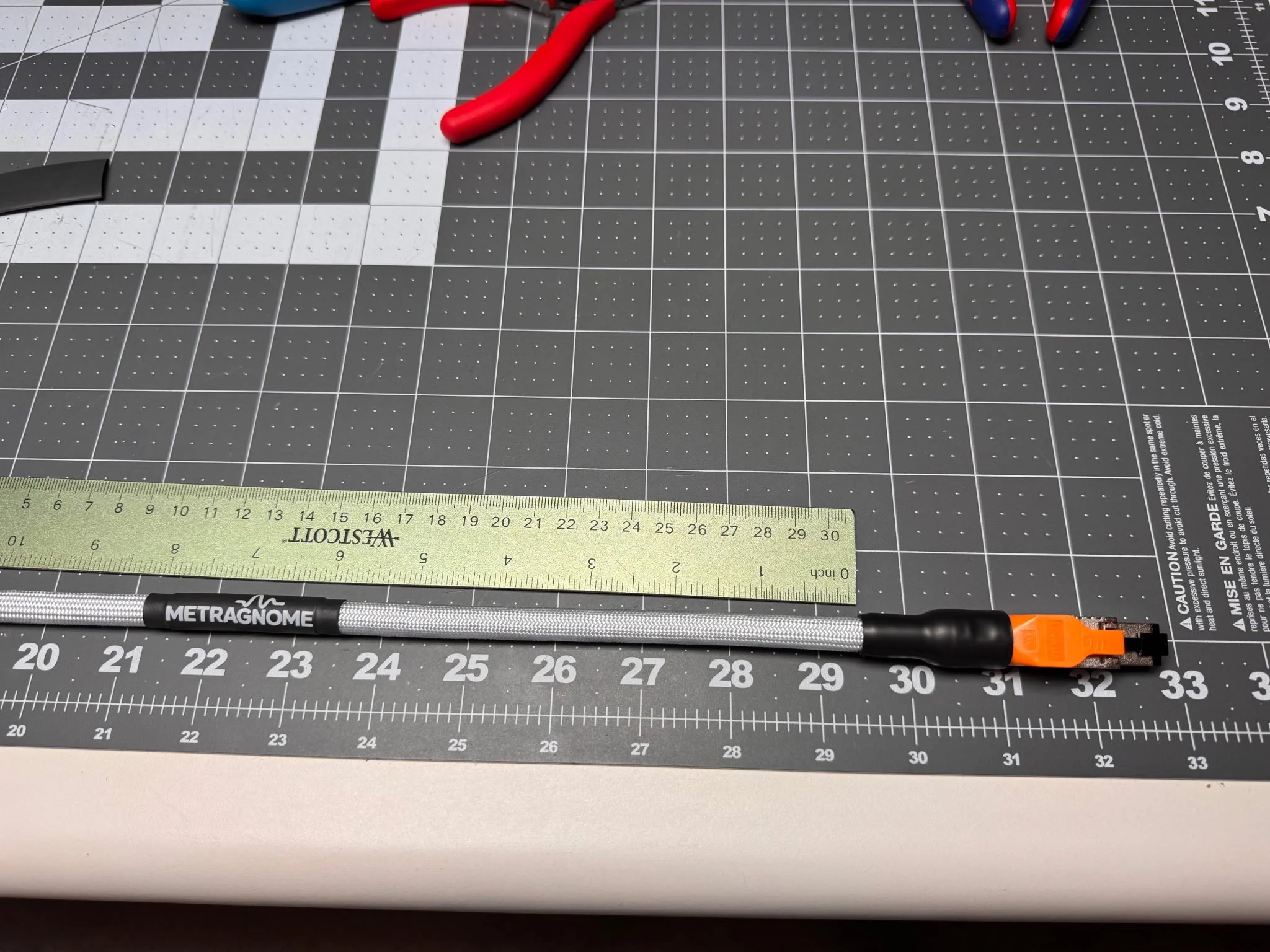 A black cutting mat with measurement markings, a yellow ruler, a black METRAGNOME marker pen, and an orange utility knife on top.