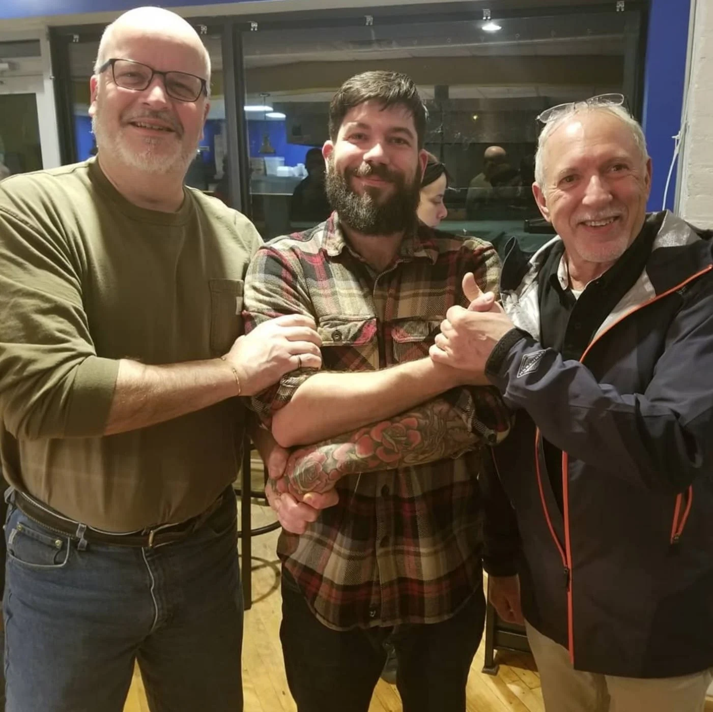 Three men smiling and holding hands together at a social gathering indoors. They are standing close to each other, with one giving a thumbs up. The background shows large windows and a dimly lit room.