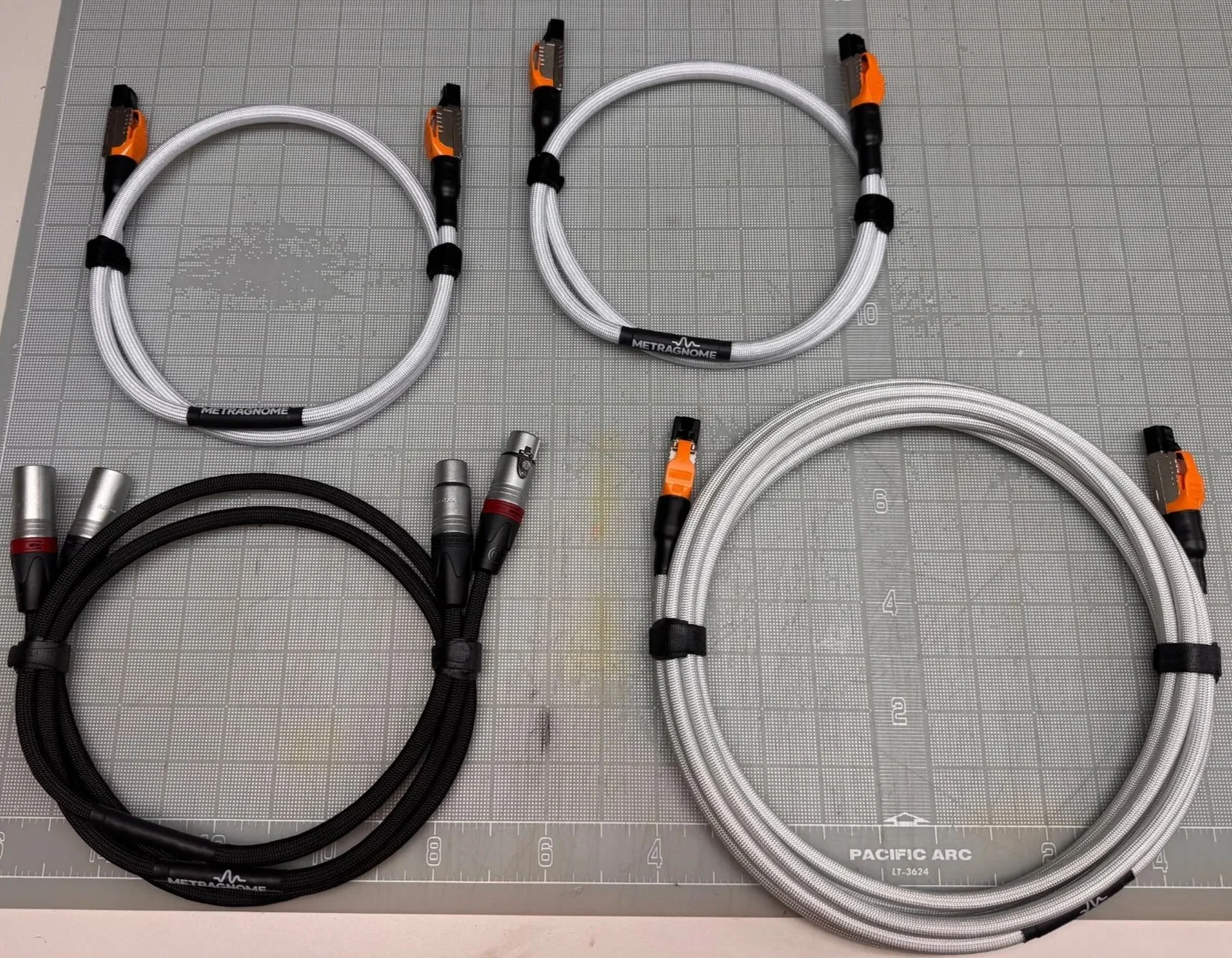 Several high-end audio or power cables with black and silver braided exteriors and orange, red, and black connectors on a gray grid work surface.