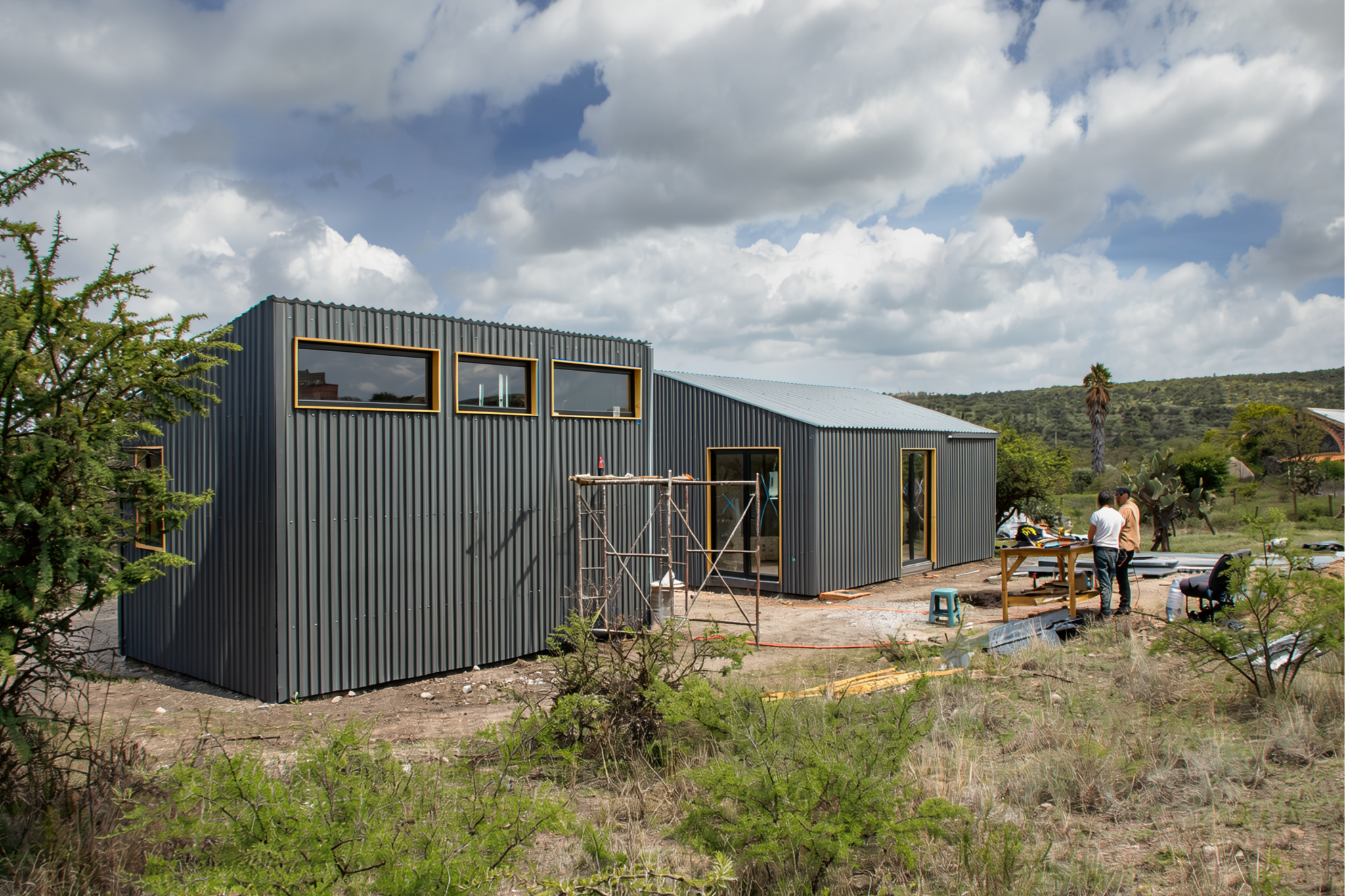 Construcción de una casa moderna con paredes de metal gris y ventanas grandes, en un entorno natural con vegetación y personas trabajando en el sitio.