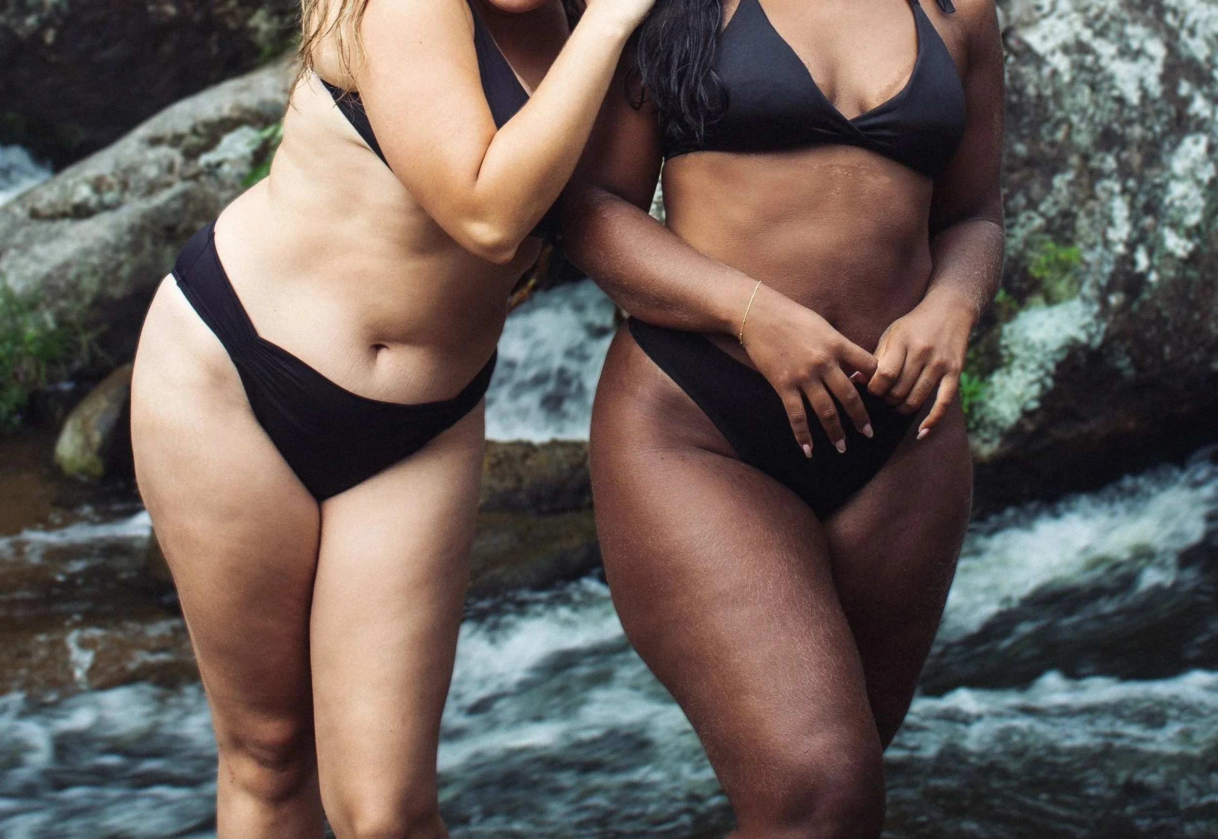 Two women in black swimsuits posing together beside a waterfall, celebrating natural beauty and body confidence.