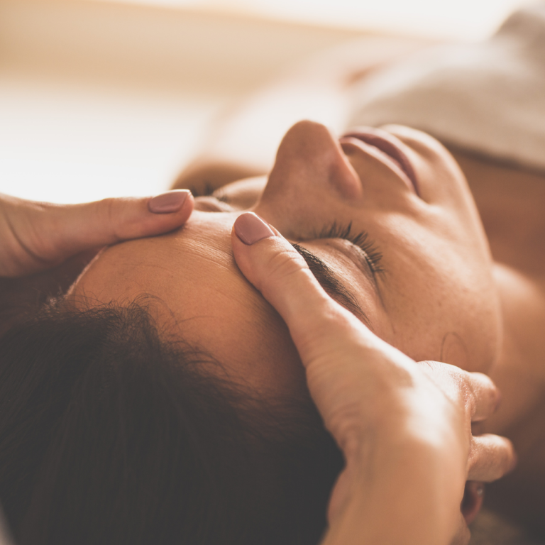Une personne reçoit un massage sur le visage, allongée sur une surface confortable, avec l'attention portée aux traits du visage, dans une ambiance relaxante.