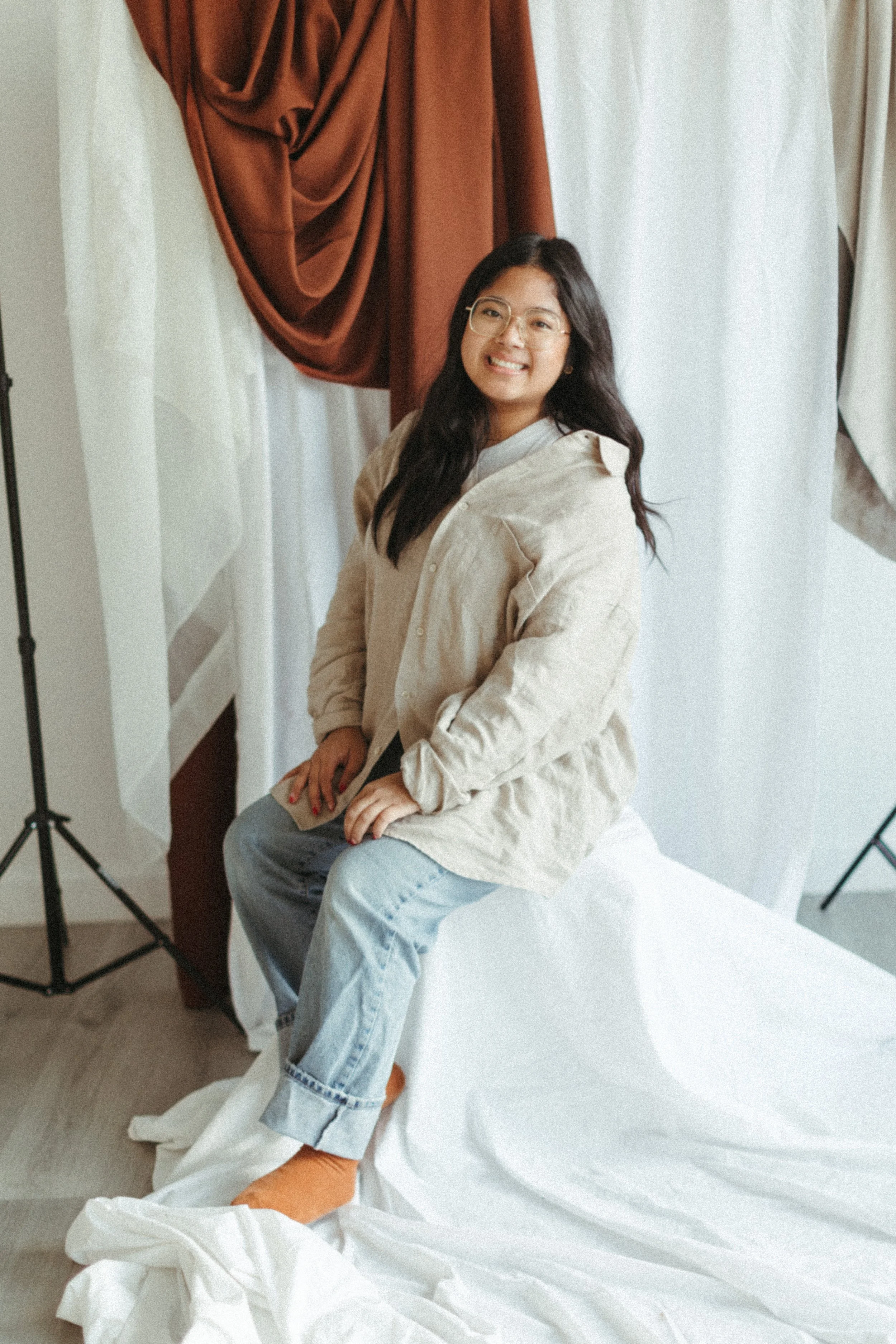 A woman with glasses sitting on a white sheet, smiling in a photography studio with brown and white curtains.