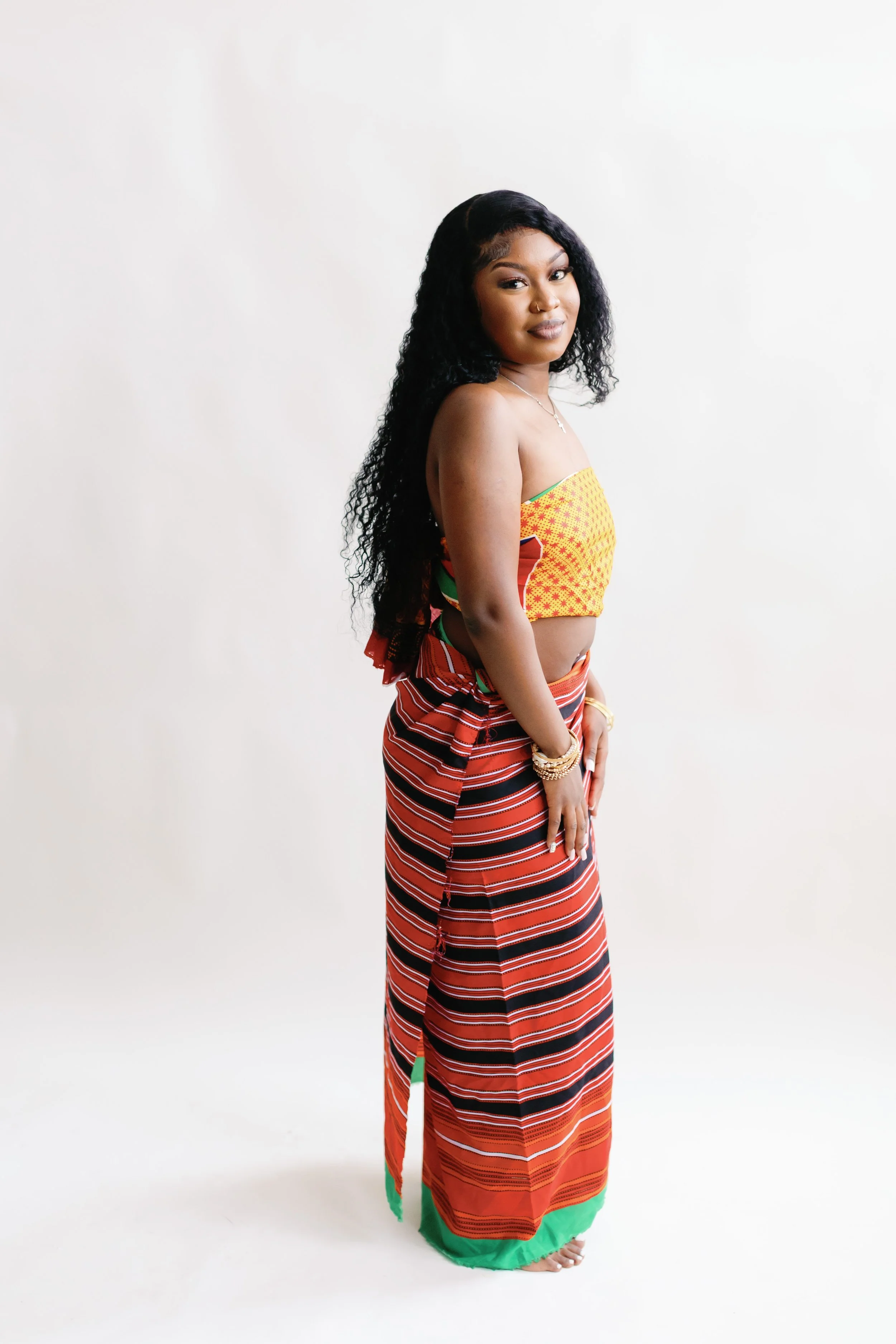 A woman with long dark curly hair wearing a colorful traditional dress with yellow, red, and green patterns, standing barefoot against a plain white background.