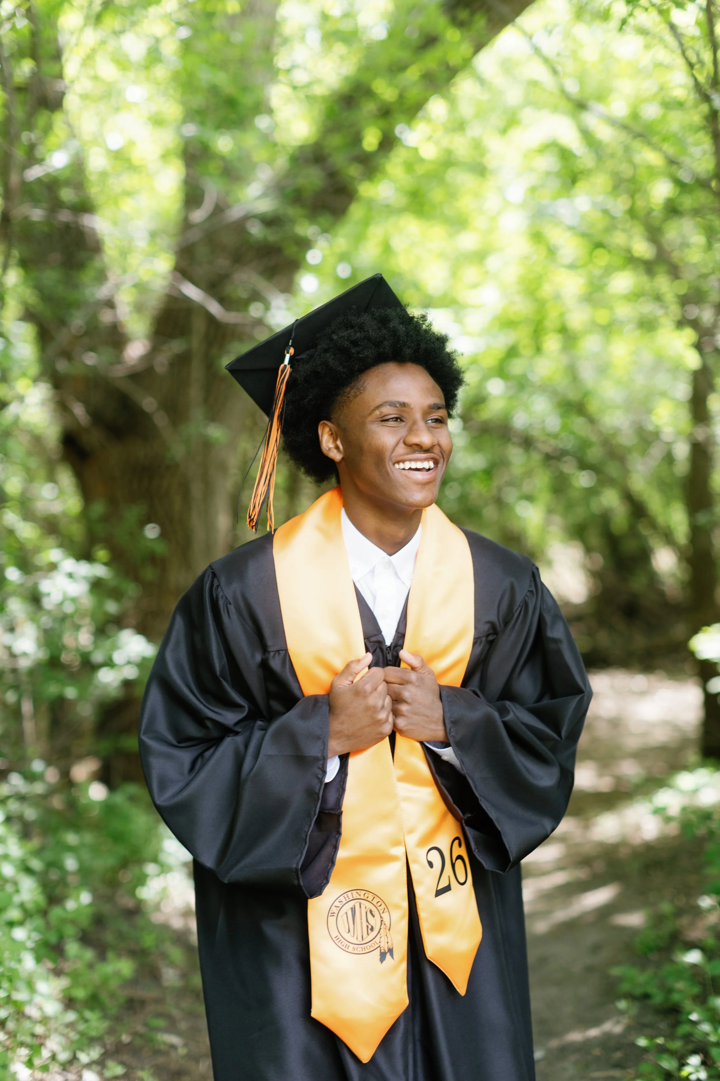 A young African American woman in graduation attire, wearing a black cap and gown with a gold stole, smiling and holding the edges of her gown, outdoors with green trees and sunlight in the background.