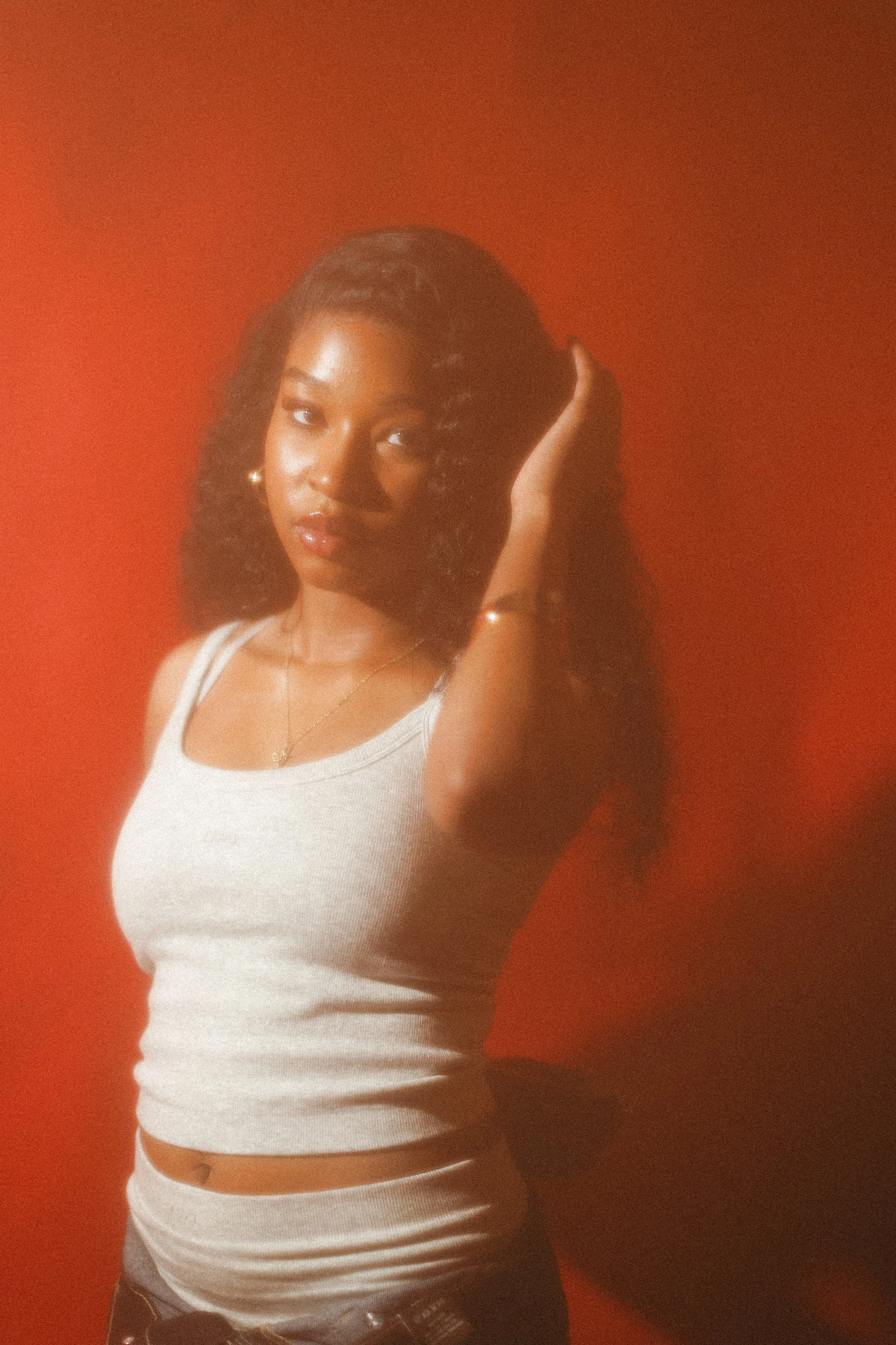 A woman with dark, wavy hair and a warm skin tone, wearing a white ribbed tank top and jewelry, standing against a vibrant orange-red background, posing with her right hand touching her hair.