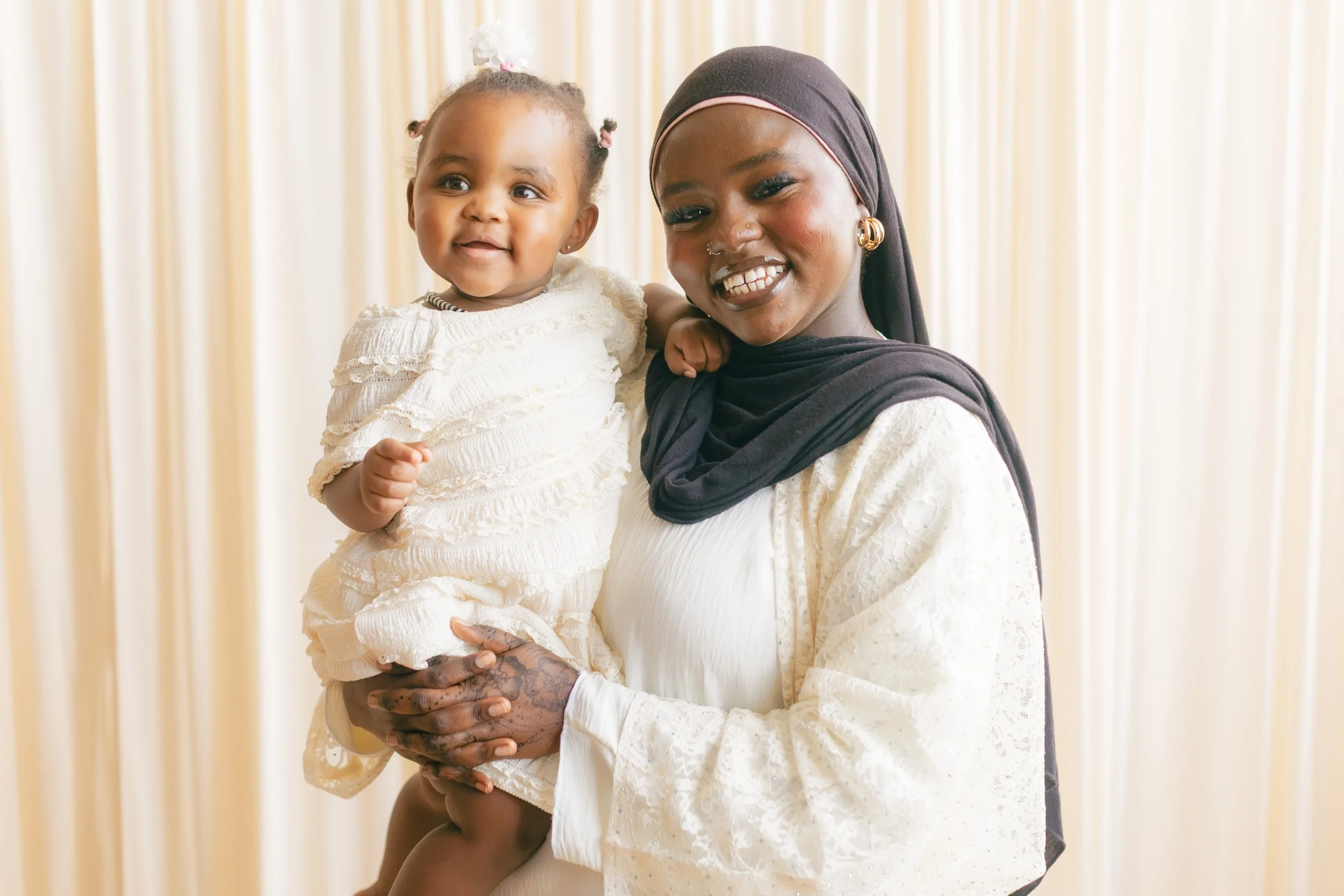 A woman wearing a black headscarf and gold earrings is smiling while holding a young girl with braided hair decorated with white flowers, both dressed in cream-colored outfits, standing in front of a beige curtain.