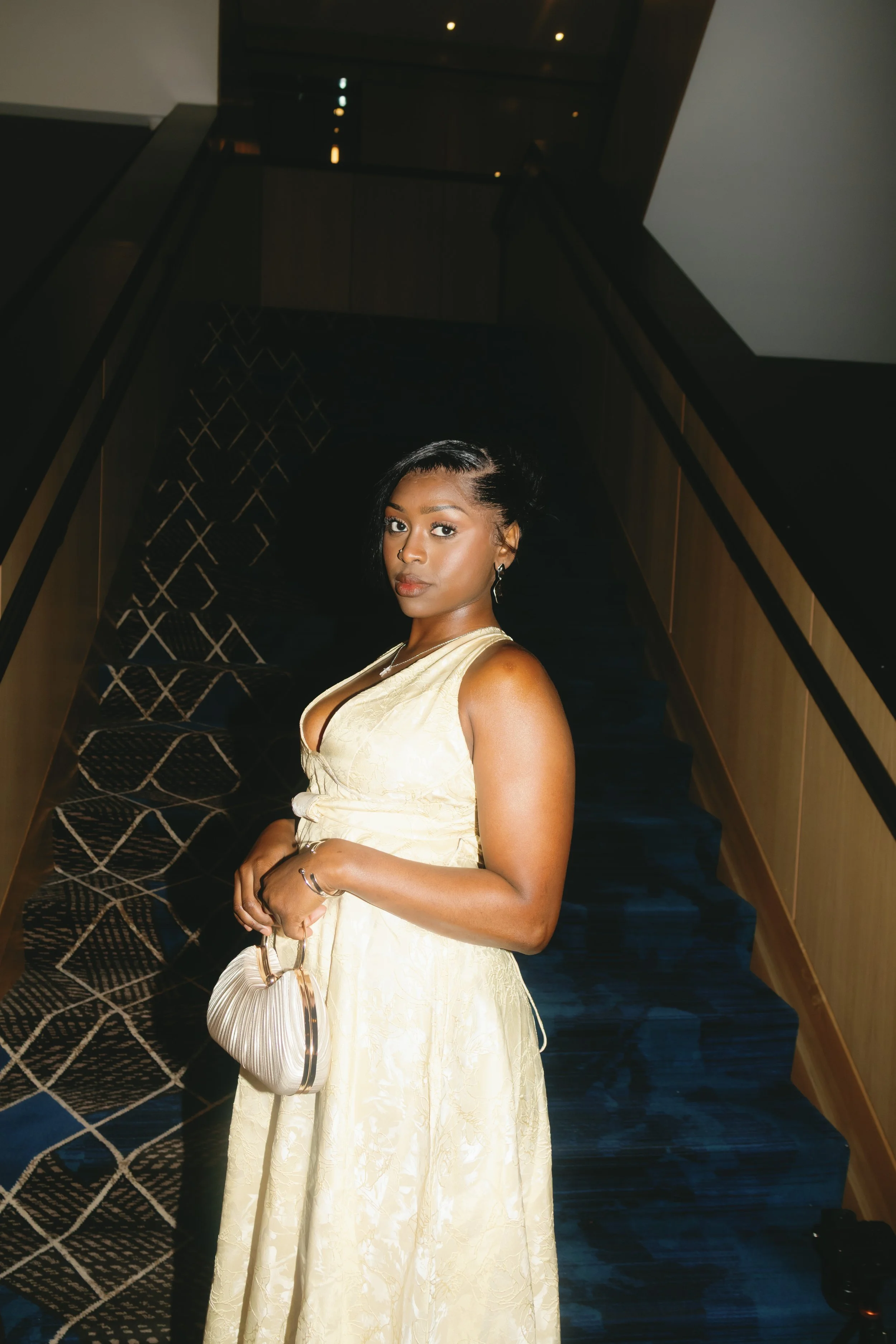 A woman in a gold dress holding a matching gold handbag, standing in a dimly lit hallway with a staircase and patterned carpet.