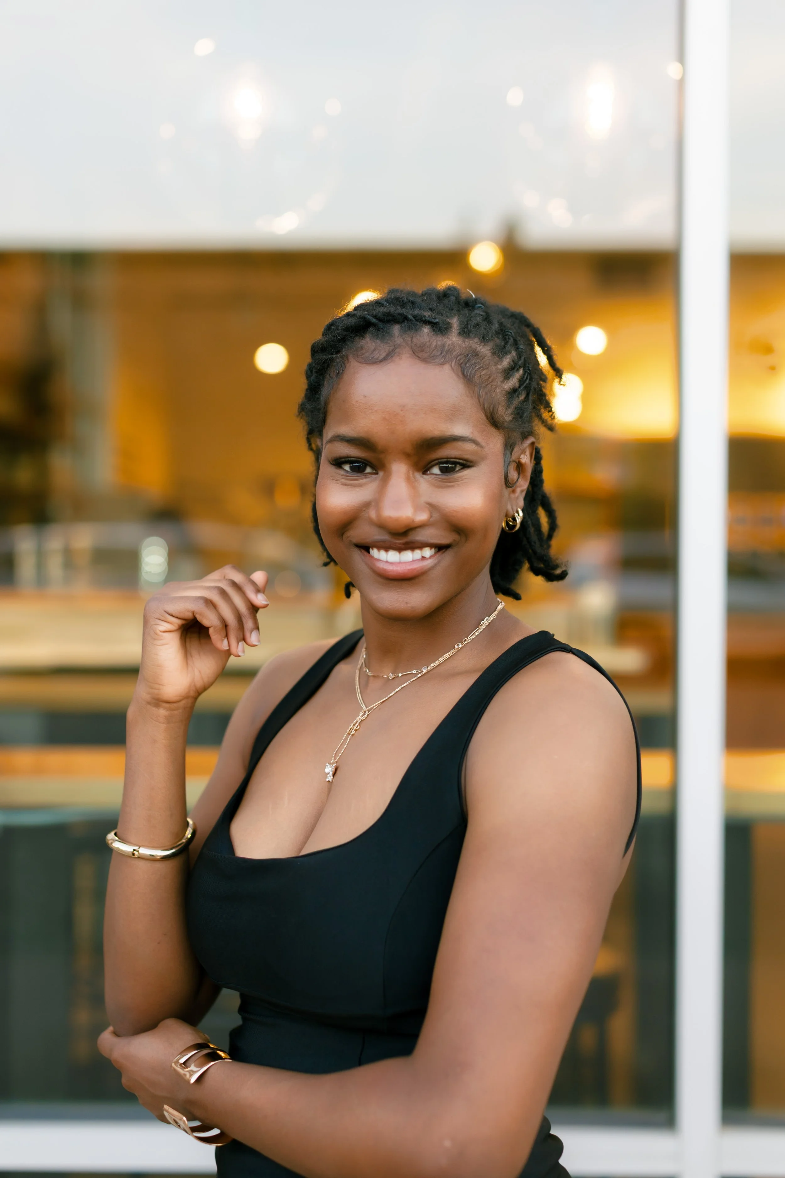 A young woman with short dreadlocks, wearing a black sleeveless top, jewelry including necklaces, earrings, and bracelets, smiling and looking at the camera indoors with warm lighting and a wooden background.