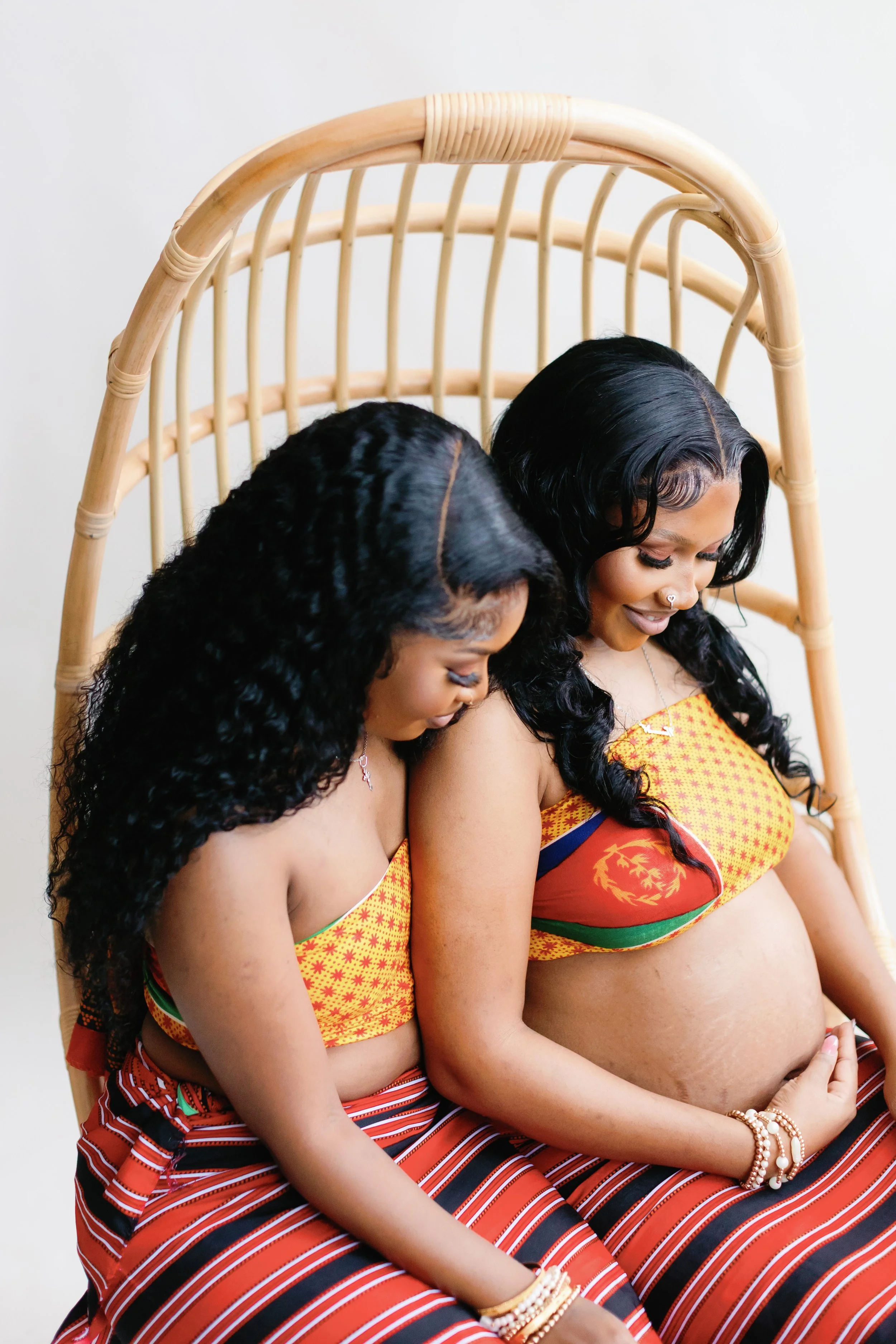 Two pregnant women with long black curly hair dressed in colorful traditional attire, sitting closely with their heads tilted down and smiling gently.