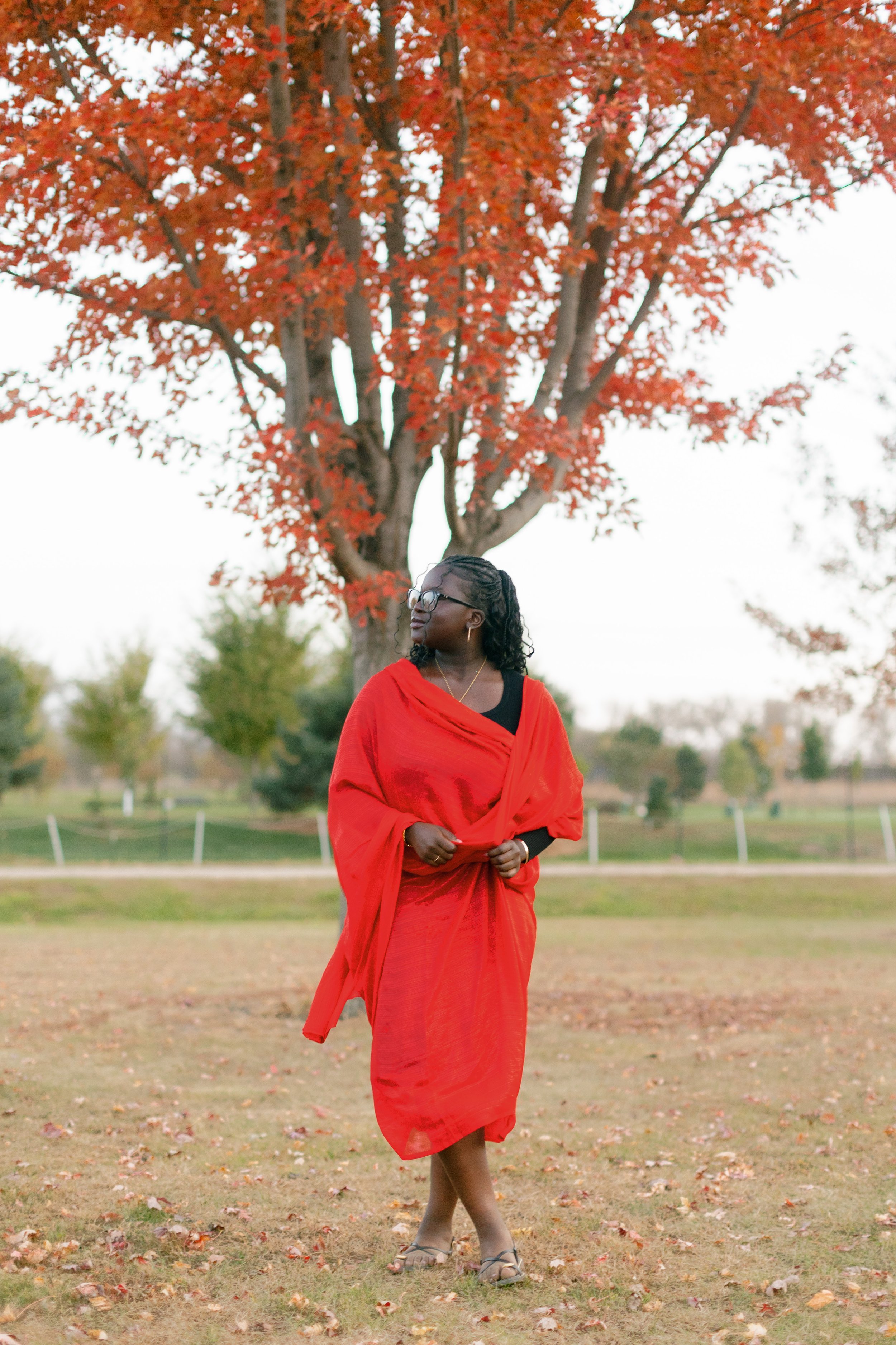 A woman dressed in a red saree stands outdoors in a park with fall foliage. She wears glasses, has curly hair, and is looking to her left. The background features a large tree with red leaves and a grassy area with fallen leaves.