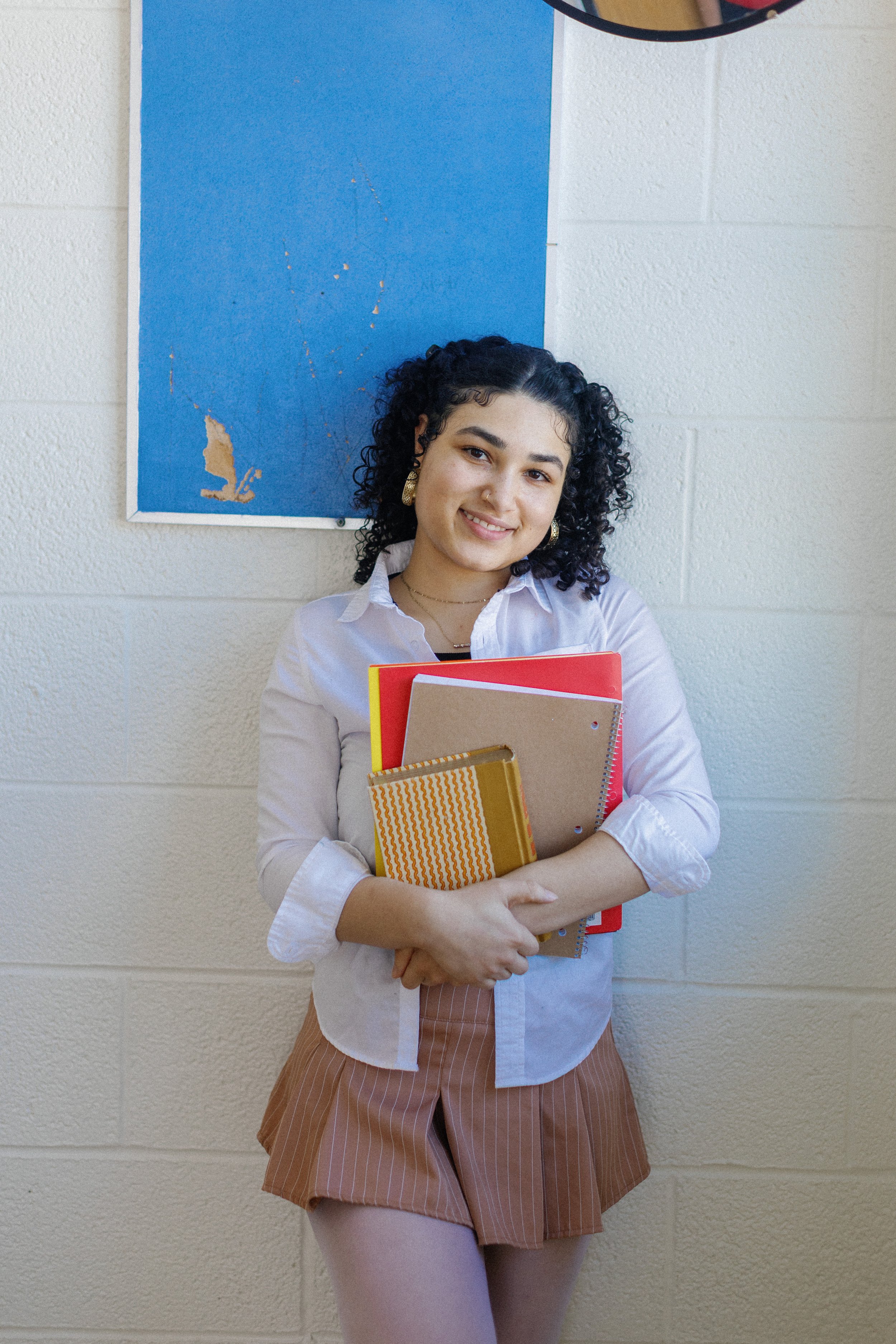 A young woman with dark curly hair, wearing a white blouse, brown striped shorts, and pink tights, stands against a light-colored cinder block wall, smiling and holding a stack of notebooks and folders.
