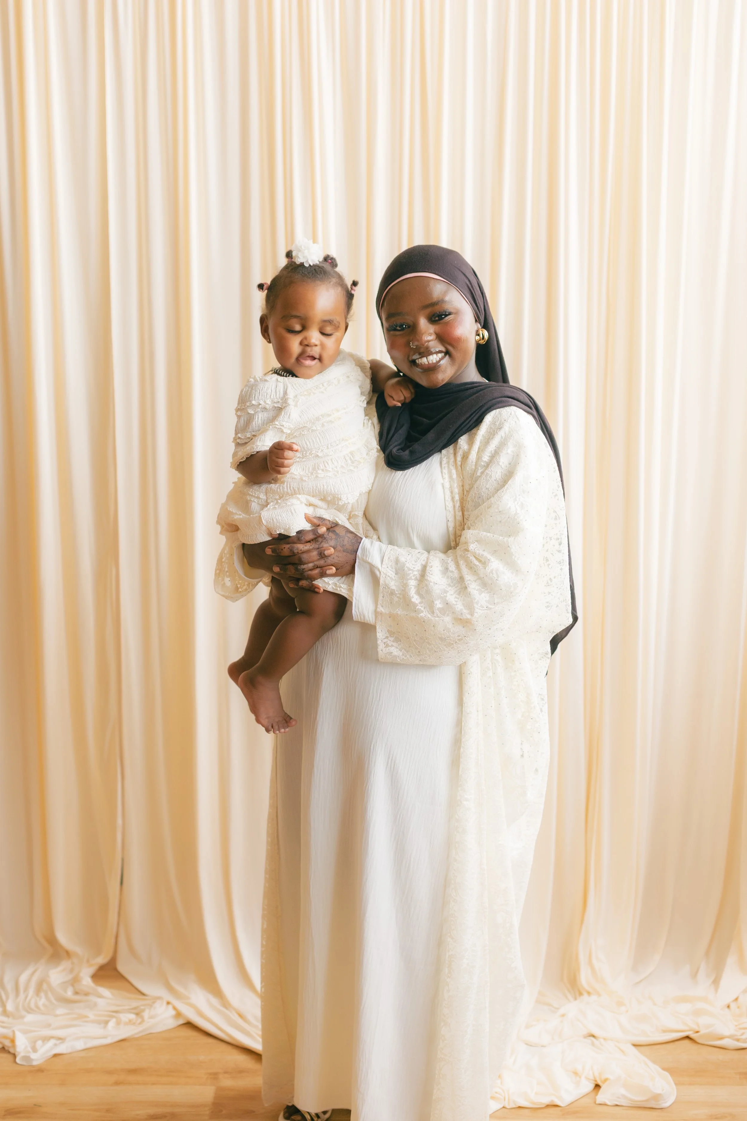 A woman wearing a black headscarf and earrings holding a young girl in a white dress in front of beige curtains.