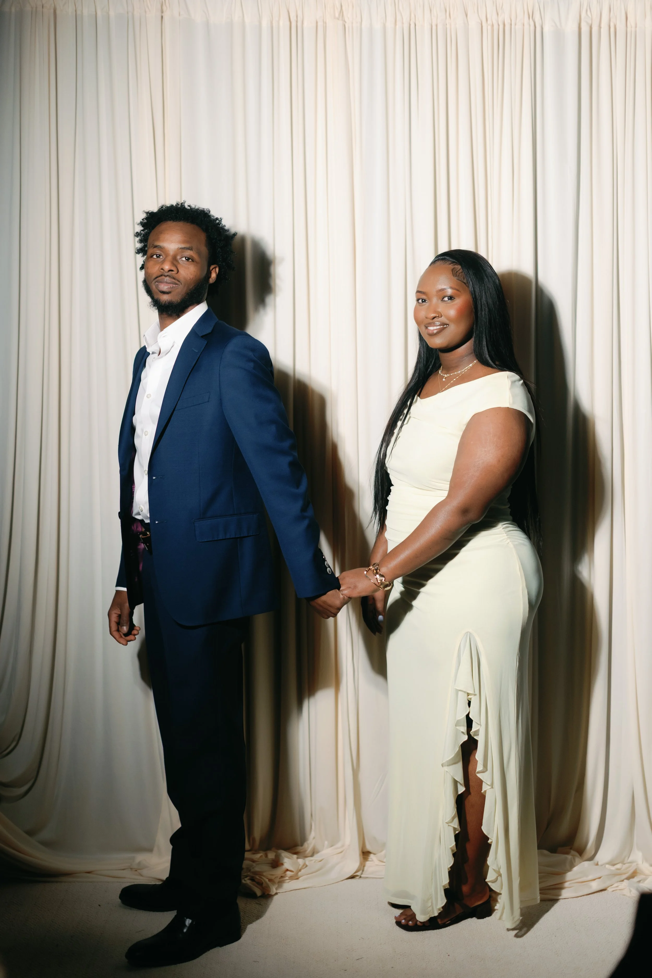 A man in a navy blazer and white shirt and a woman in a white gown holding hands and standing in front of a cream-colored curtain.