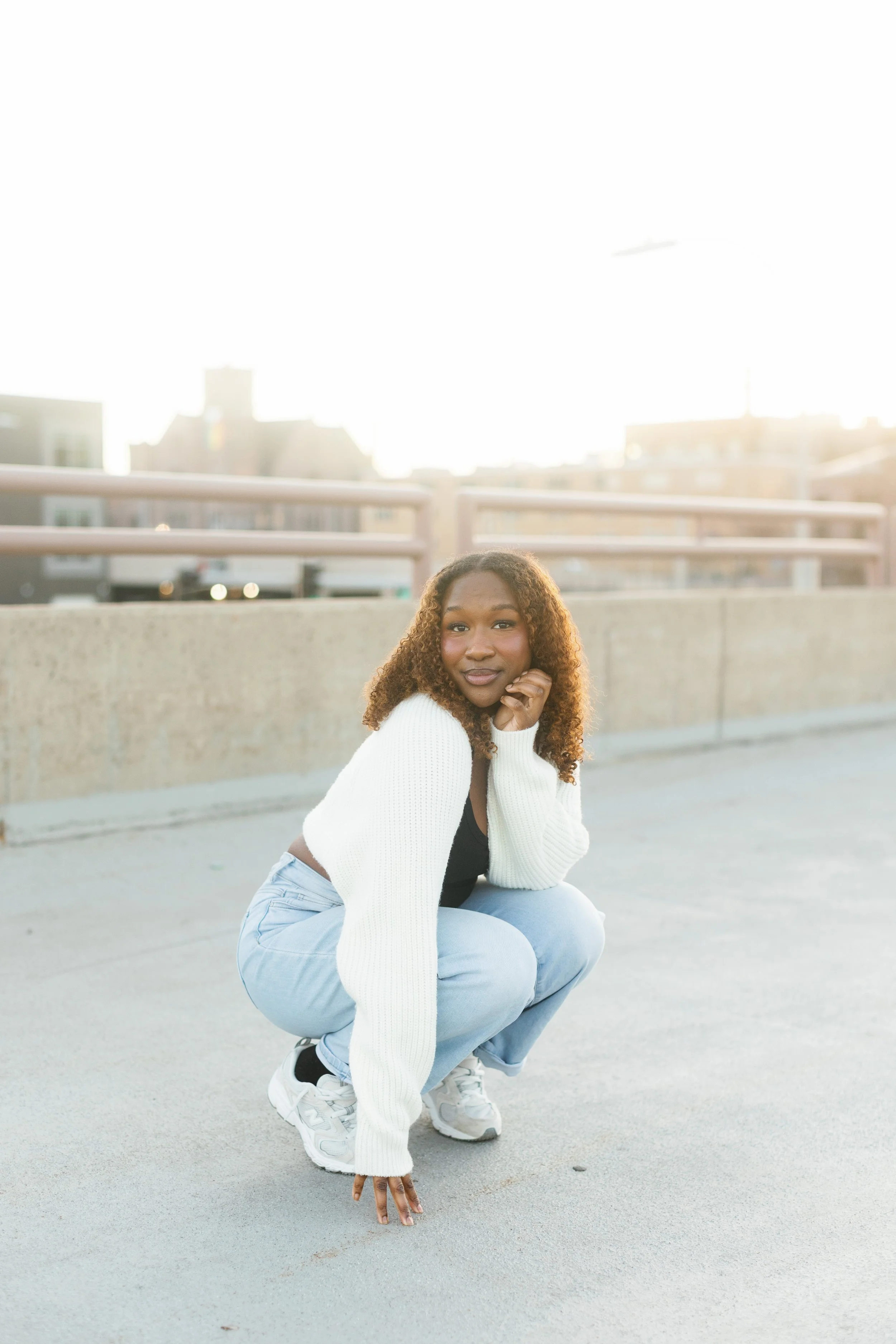 A young woman with curly hair squatting on an outdoor rooftop or parking lot, with city buildings in the background, wearing a white sweater, black top, light blue jeans, and white sneakers, looking confidently at the camera.