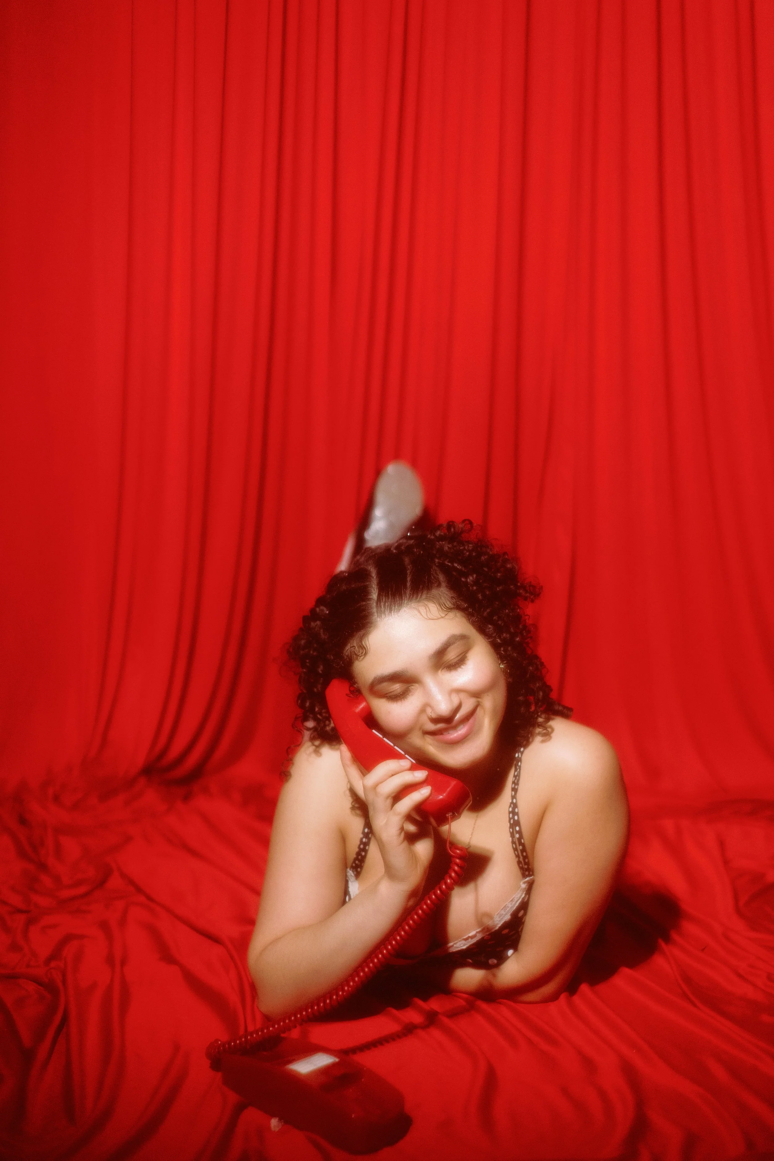 A woman with curly dark hair, wearing a spaghetti strap top, smiling while holding a red telephone receiver to her ear against a red curtain backdrop.
