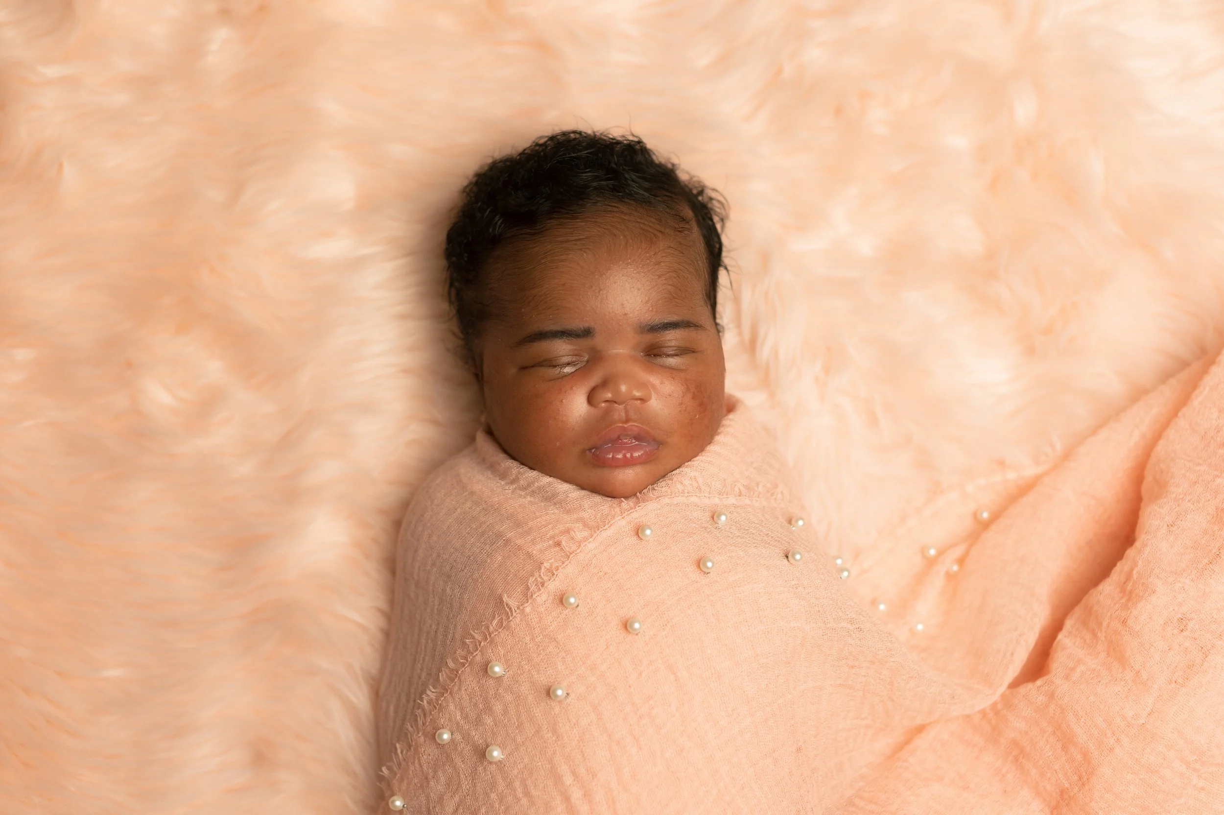 A young child with curly black hair and medium brown skin sleeping peacefully on a soft, peach-colored blanket with pearl embellishments.