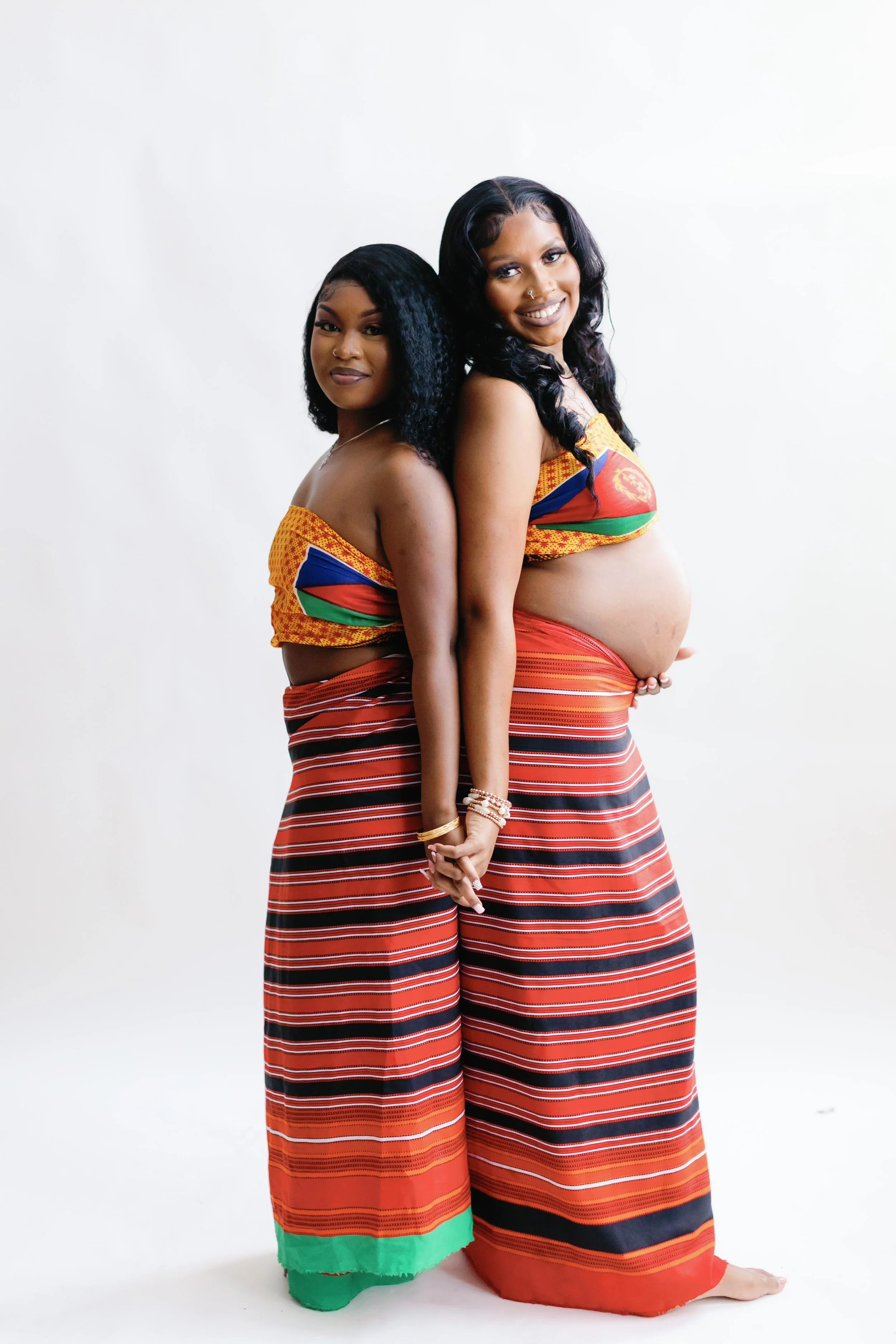 Two women with dark hair wearing matching colorful traditional outfits standing back to back against a plain white background.