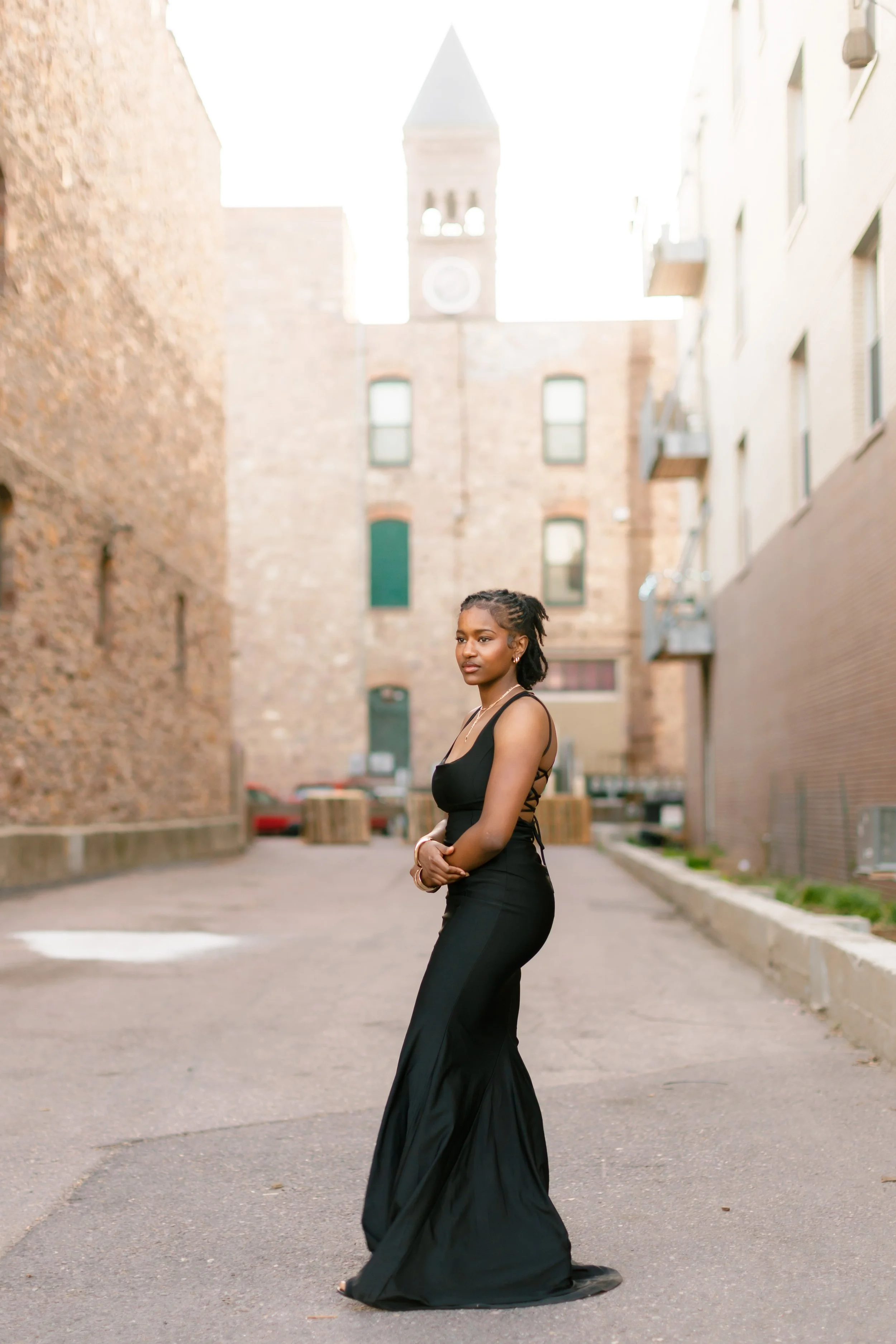 A woman in a black dress standing on a city street with old brick buildings and a clock tower in the background.