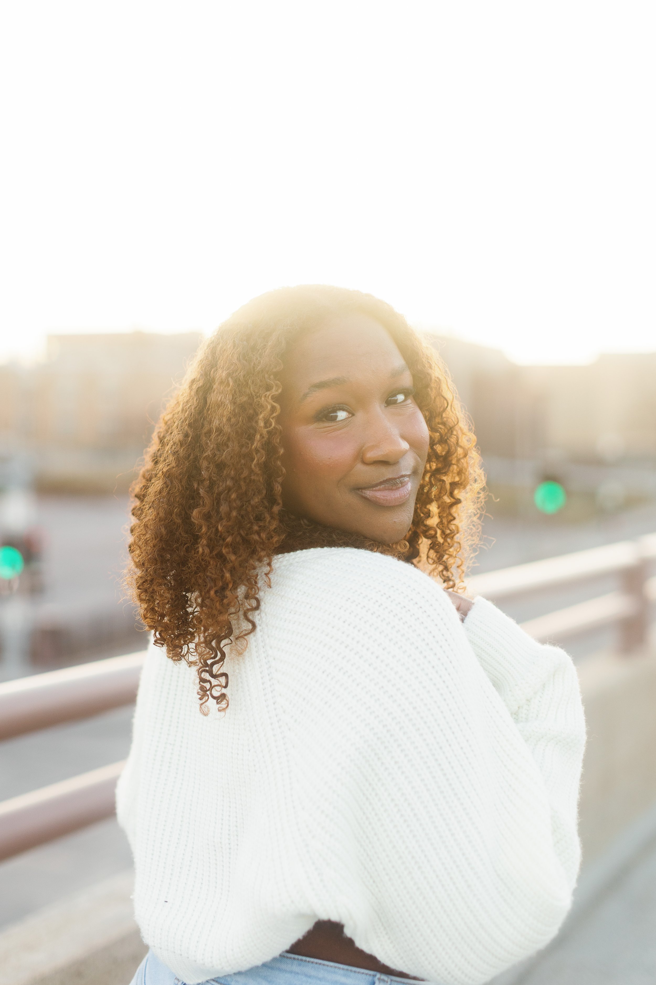 A woman with curly brown hair and light brown skin standing outdoors on a bridge at sunset, wearing a white sweater and smiling.