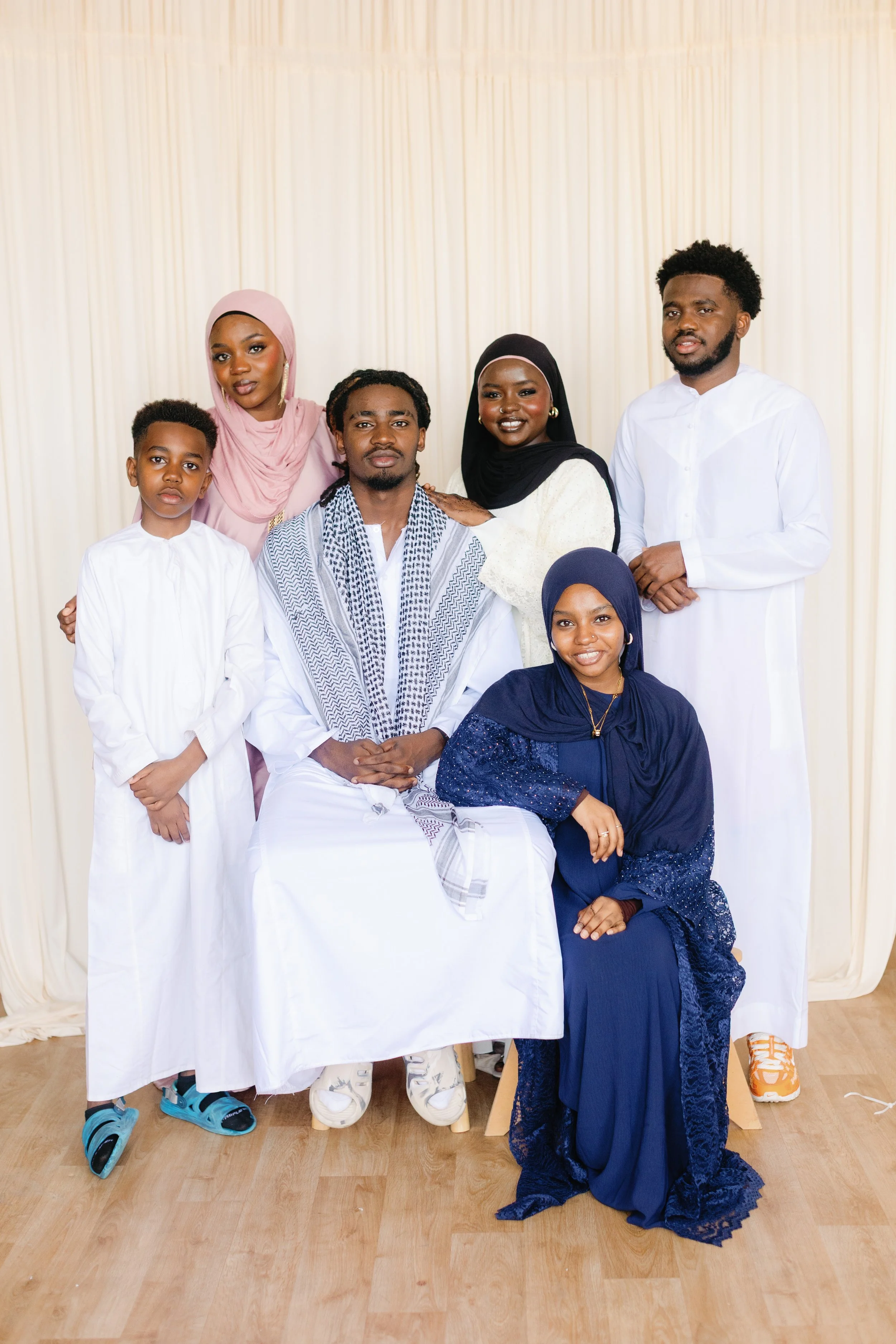 Group of seven diverse people, including men, women, and children, dressed in traditional and modern clothing, posing together in front of a cream-colored curtain indoors.