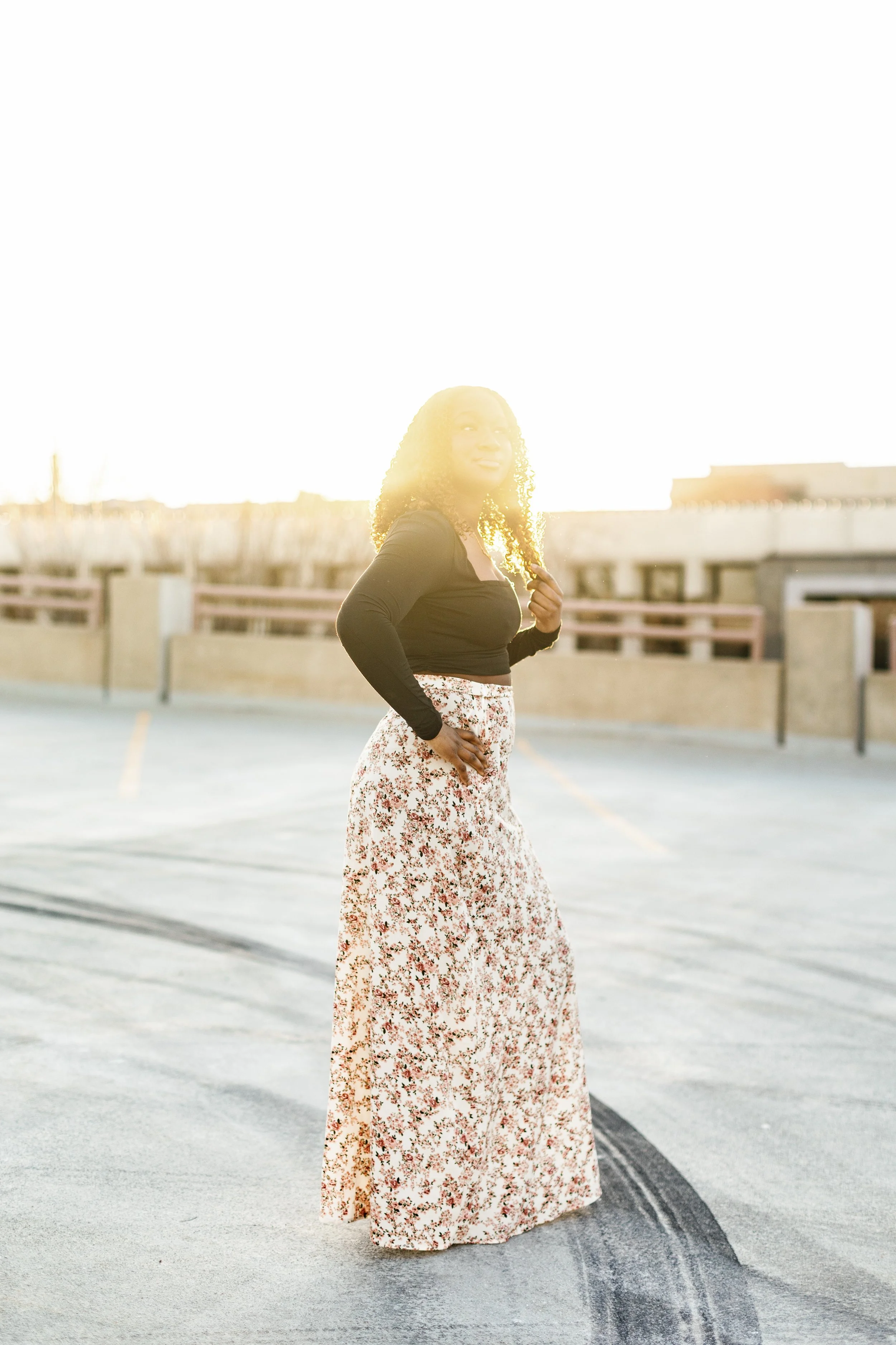 A woman standing outdoors in the late afternoon sunlight, wearing a black long-sleeve top and a long floral skirt, with her right hand on her hip and hair styled in tight curls.
