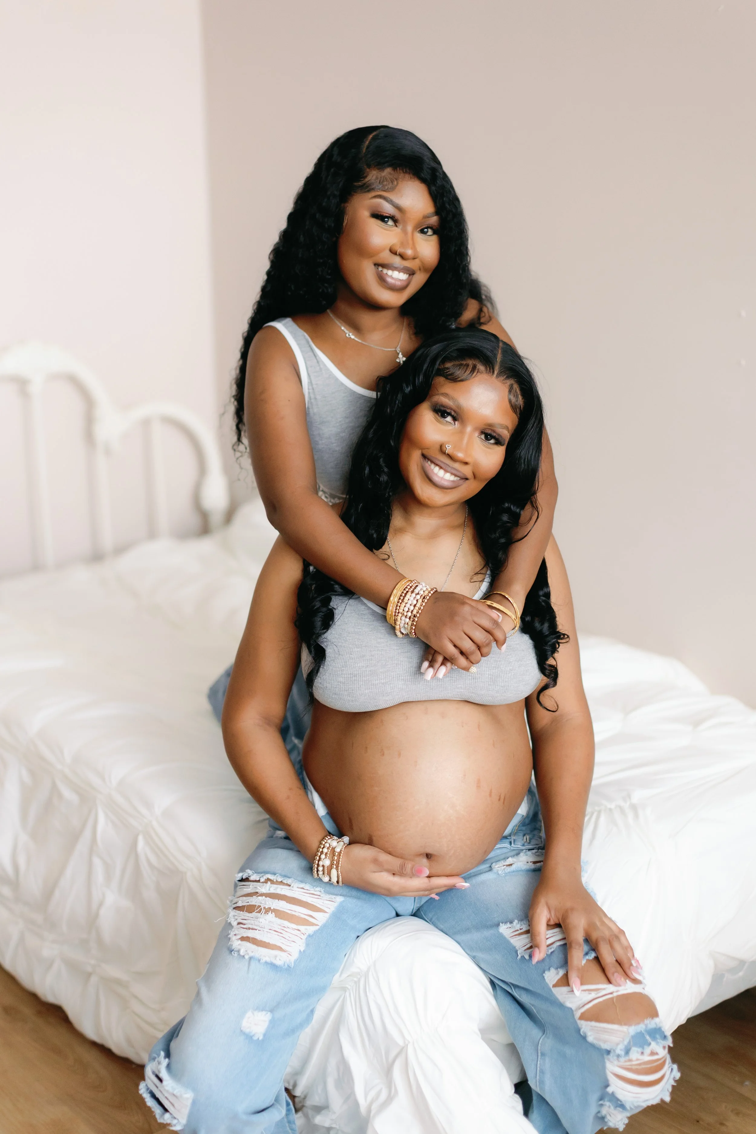 Two women, one pregnant, smiling and hugging in a bedroom with a white bed, dressed in casual clothes and jewelry.