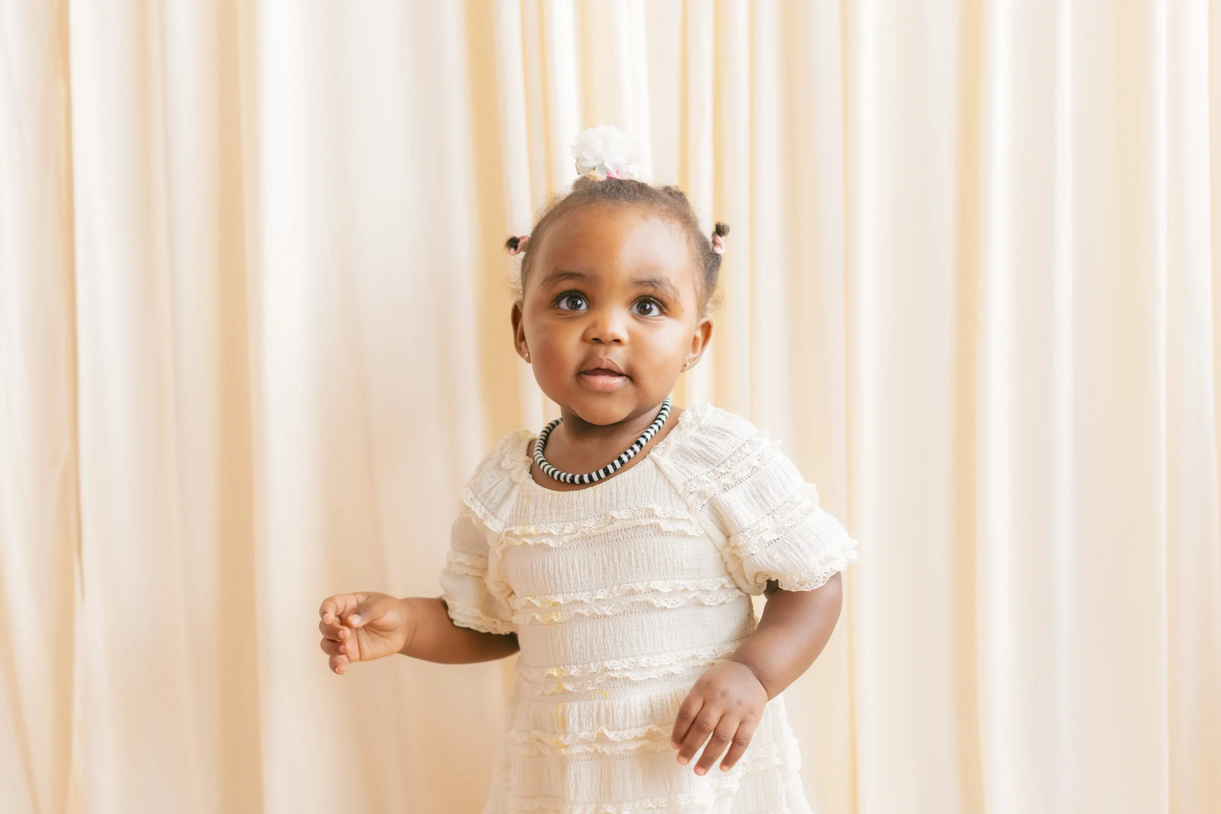 Young girl with braided hair and a white dress standing in front of a cream-colored curtain.