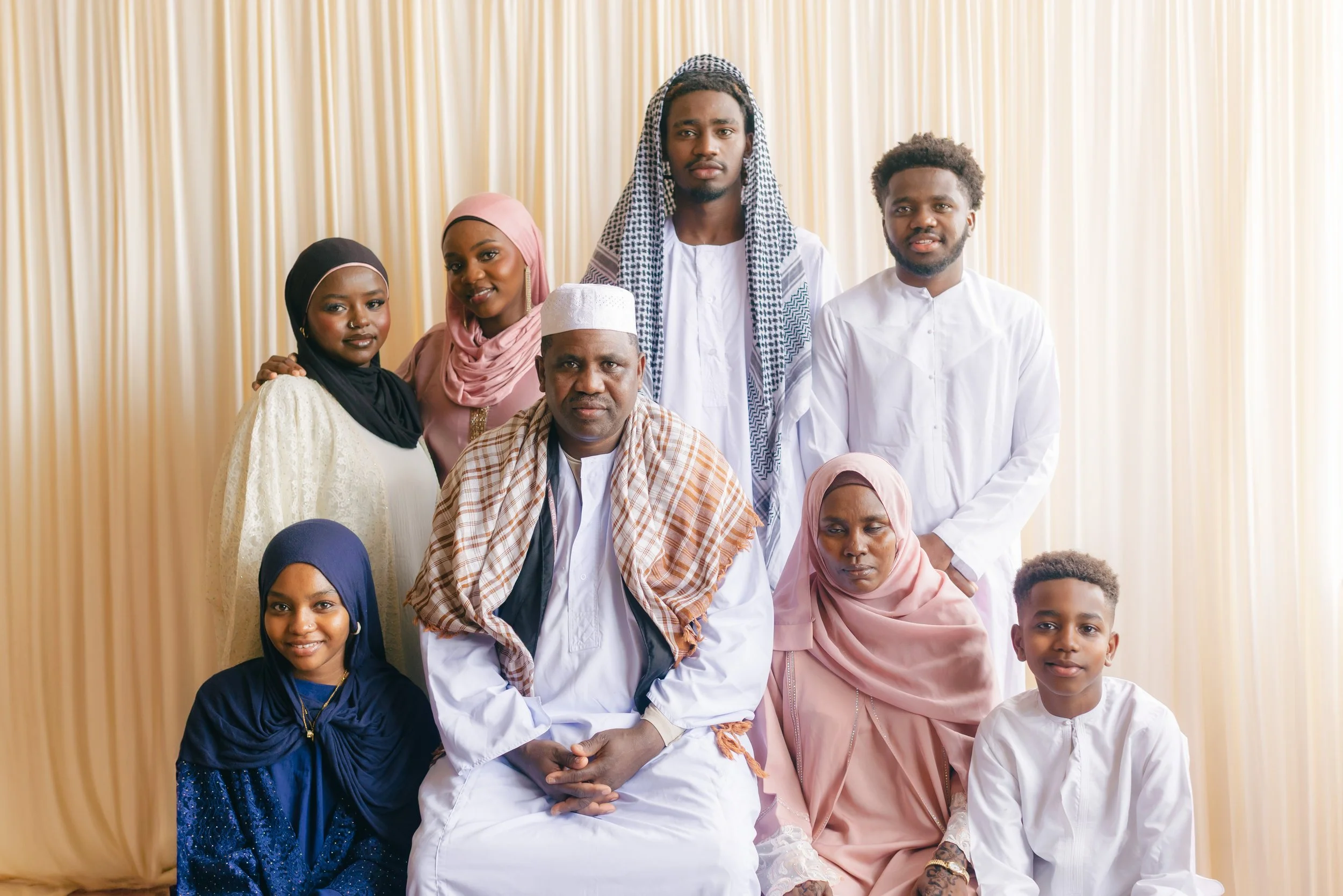 A group of diverse people dressed in traditional and casual clothing, posing in front of a cream-colored curtain.