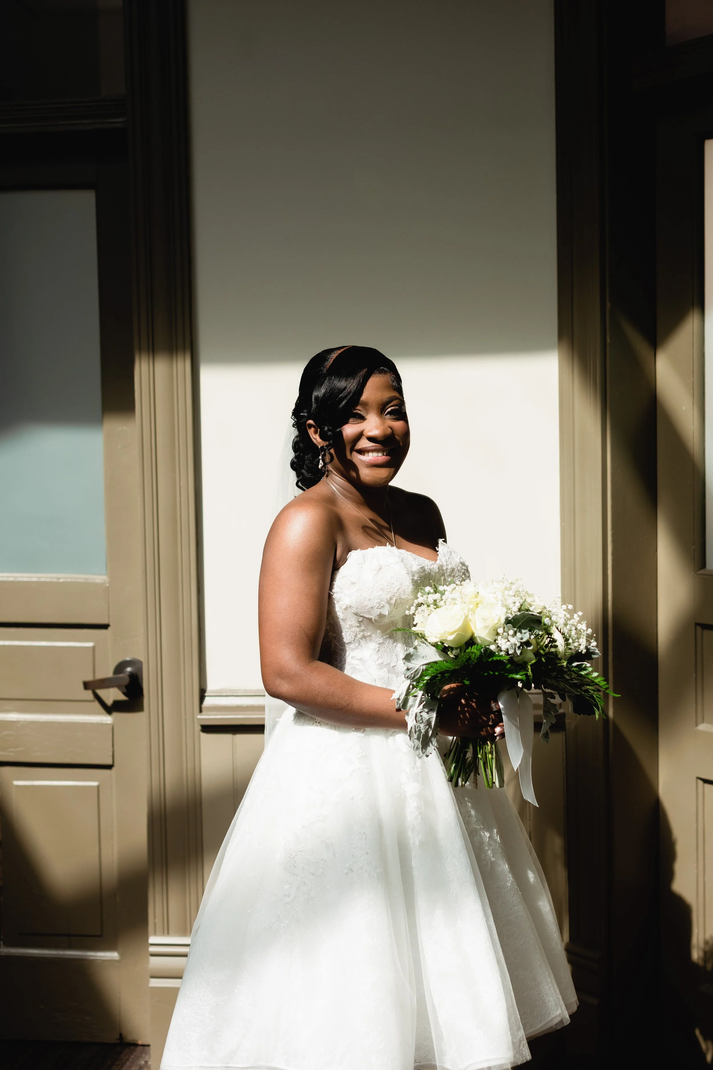 A smiling bride in a white wedding dress holding a bouquet of white roses and greenery, standing in a doorway with sunlight creating shadows on the wall behind her.