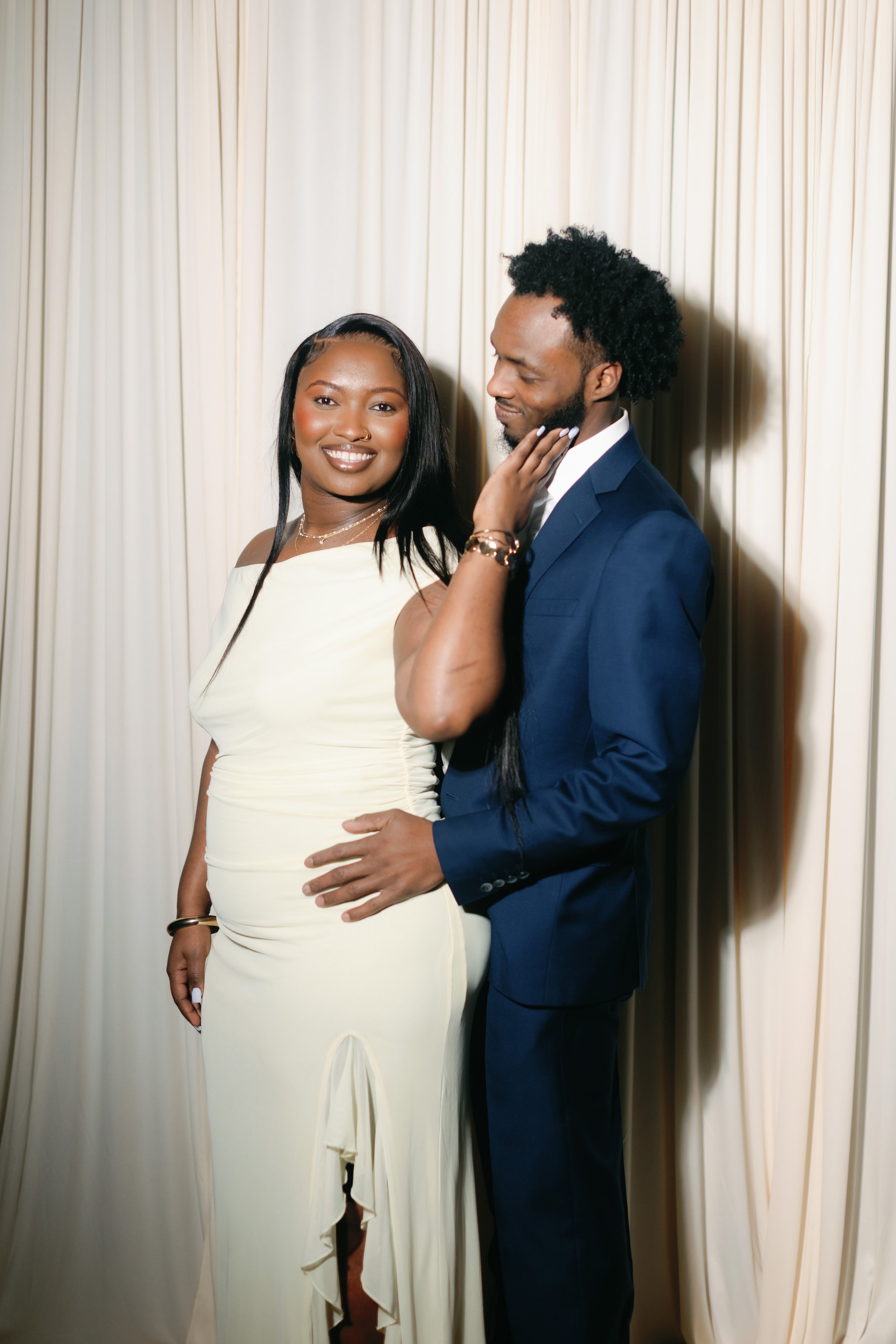 A woman in a white dress smiling at the camera while a man in a blue suit touches her face gently, standing in front of cream-colored curtains.
