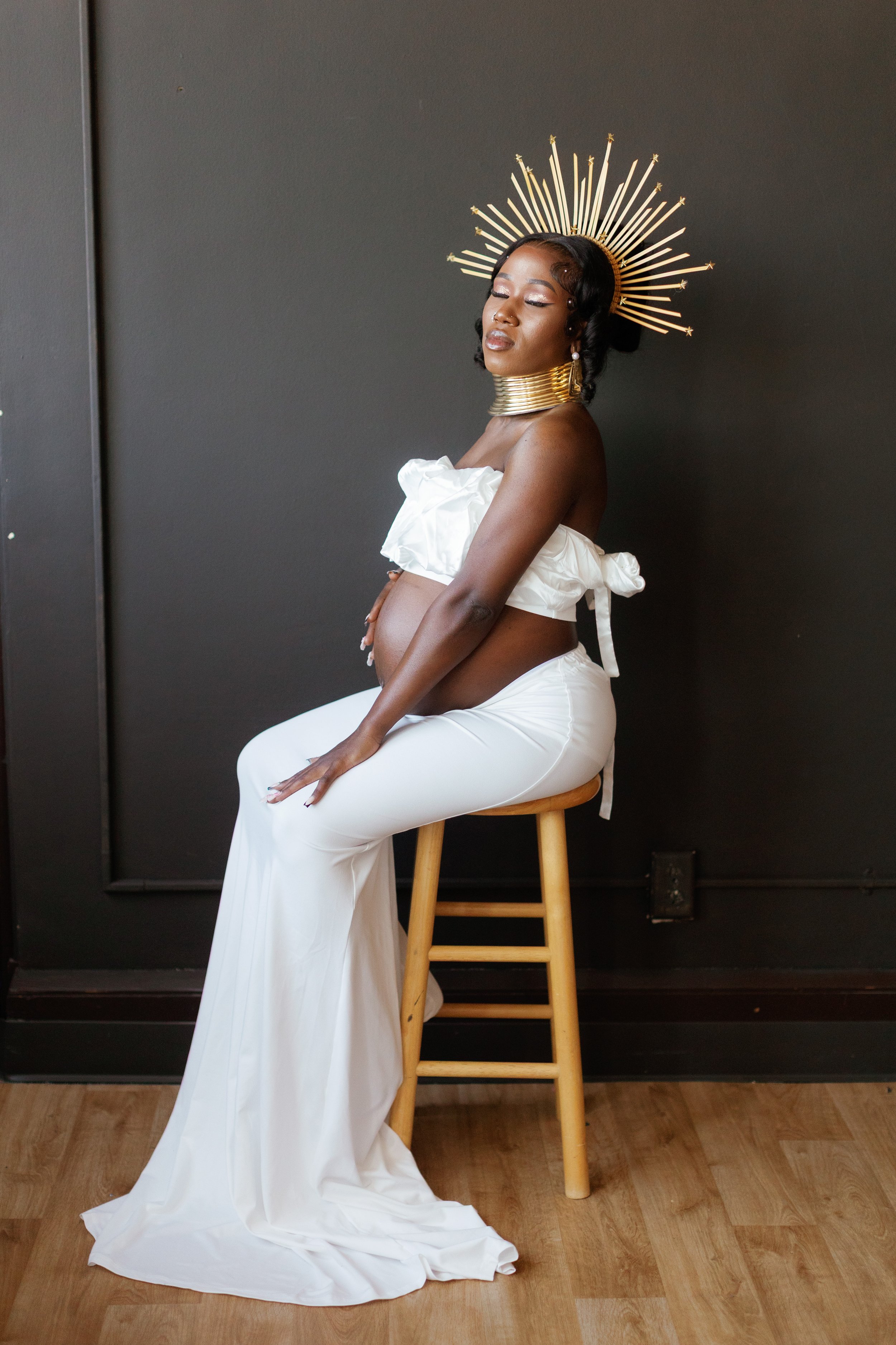 A woman dressed in white with a gold headpiece, gold choker, and earrings, sitting on a wooden stool against a black wall.