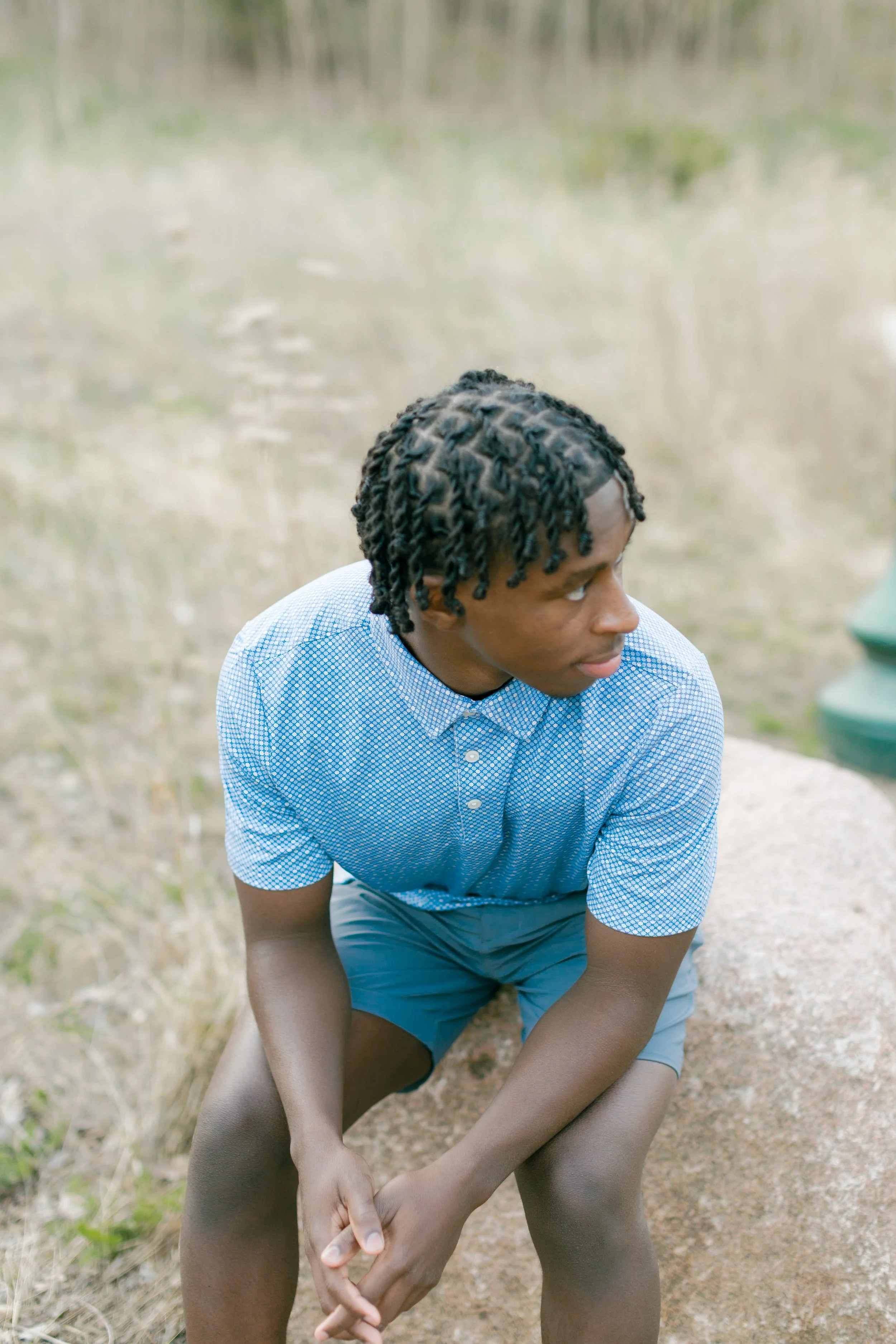 A young man with medium skin tone, wearing a light blue and white checkered shirt and gray shorts, kneeling outdoors on dirt ground with grassy background, looking to his right.