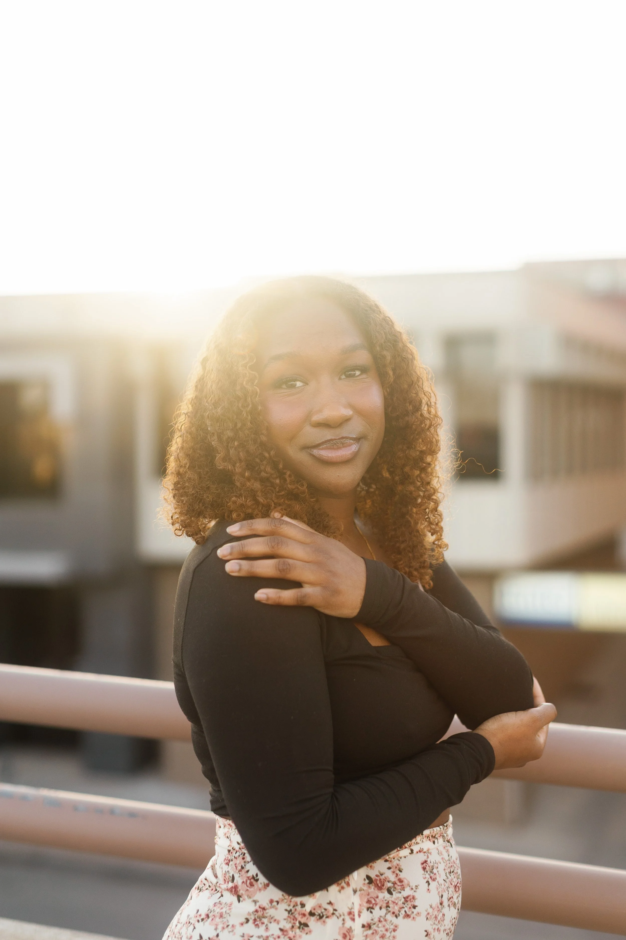 A woman with curly hair wearing a black top and floral skirt standing outdoors during sunset on a balcony with buildings in the background.