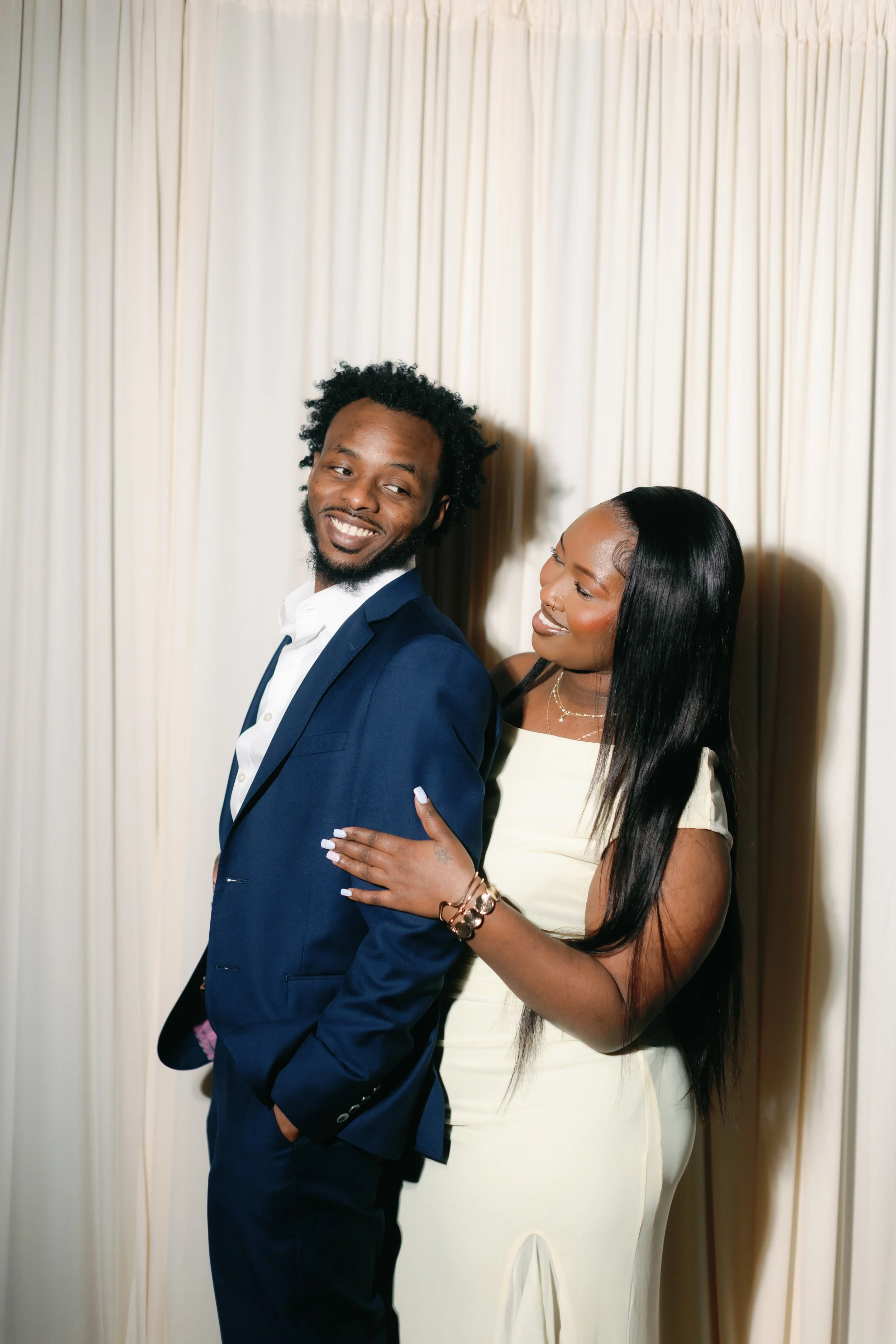 A man in a dark blue suit and woman in a cream dress standing close together in front of cream-colored curtains, smiling warmly at each other.